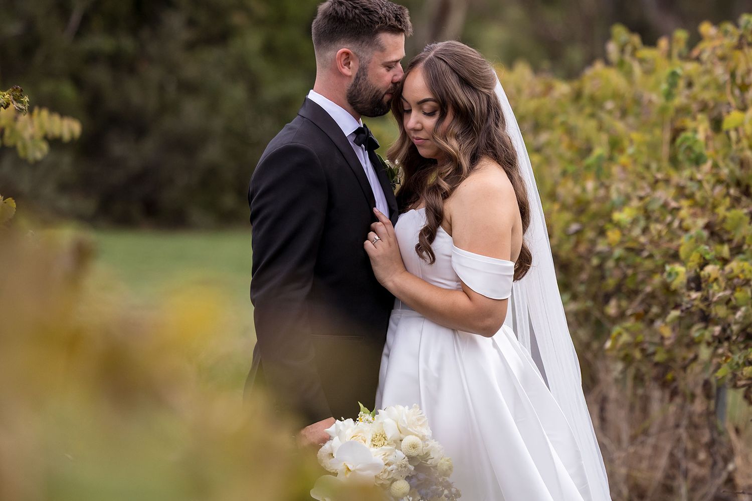 Newlywed couple embraces with white bridal bouquet among autumn foliage of the grape vines in romantic wedding moment at Brown Brothers Wine