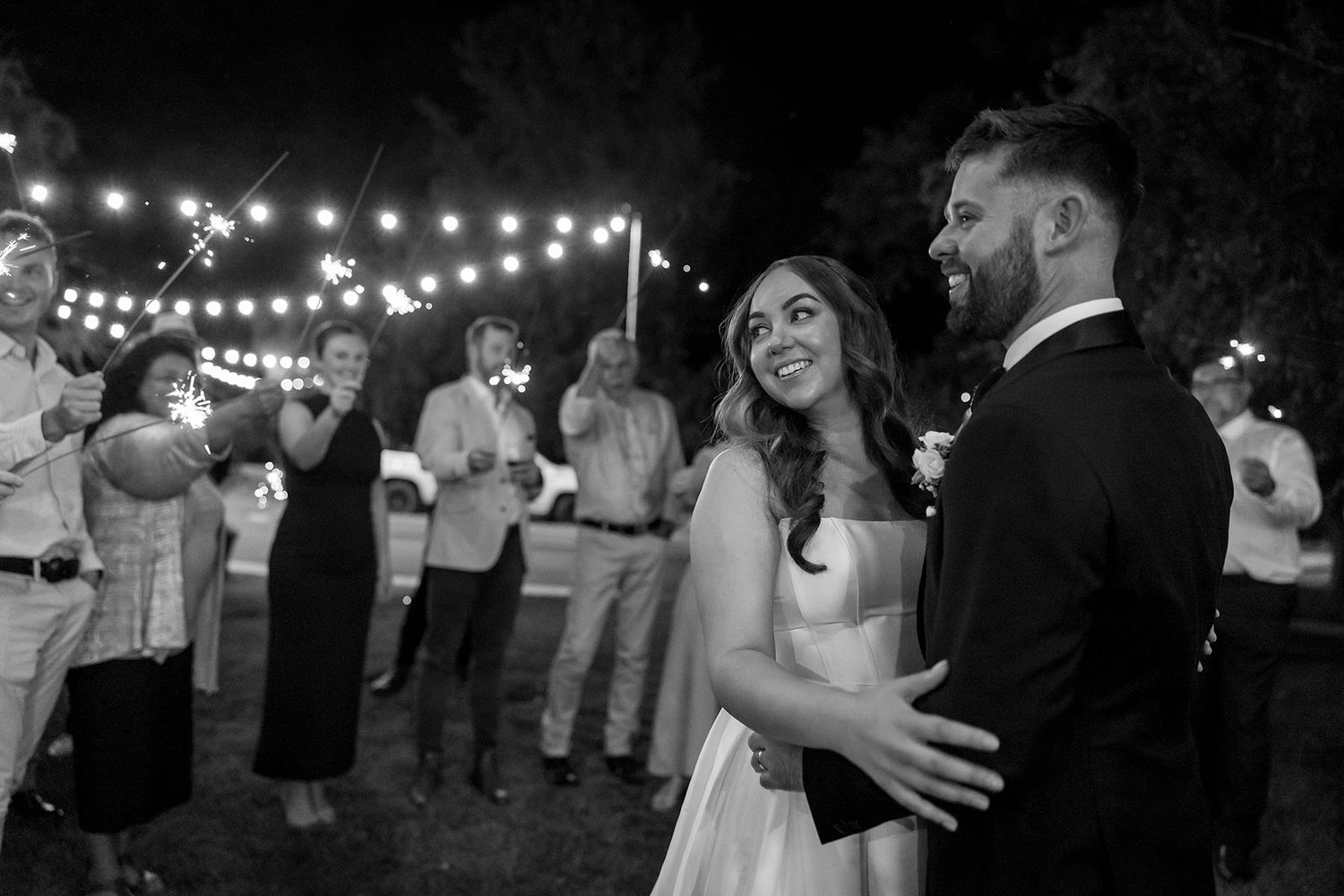 A couple shares their first dance under string lights at an outdoor evening wedding reception while guests look on at Brown Brothers Winery.