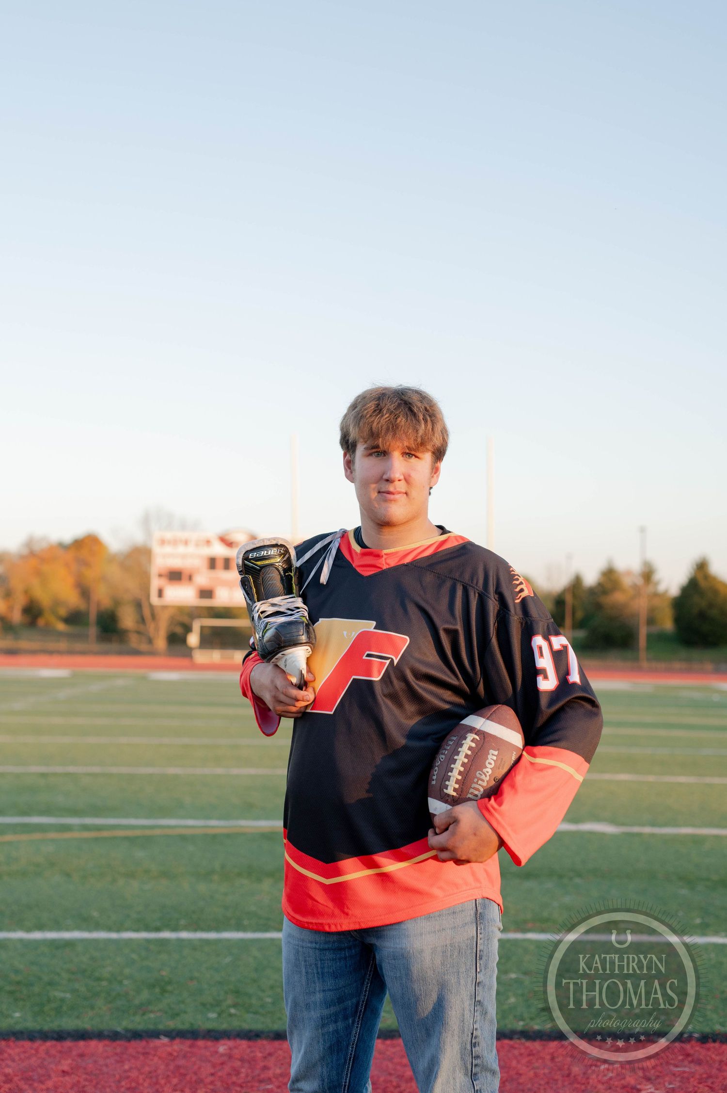Multi-sport athlete holds football and ice skates on field during golden hour.