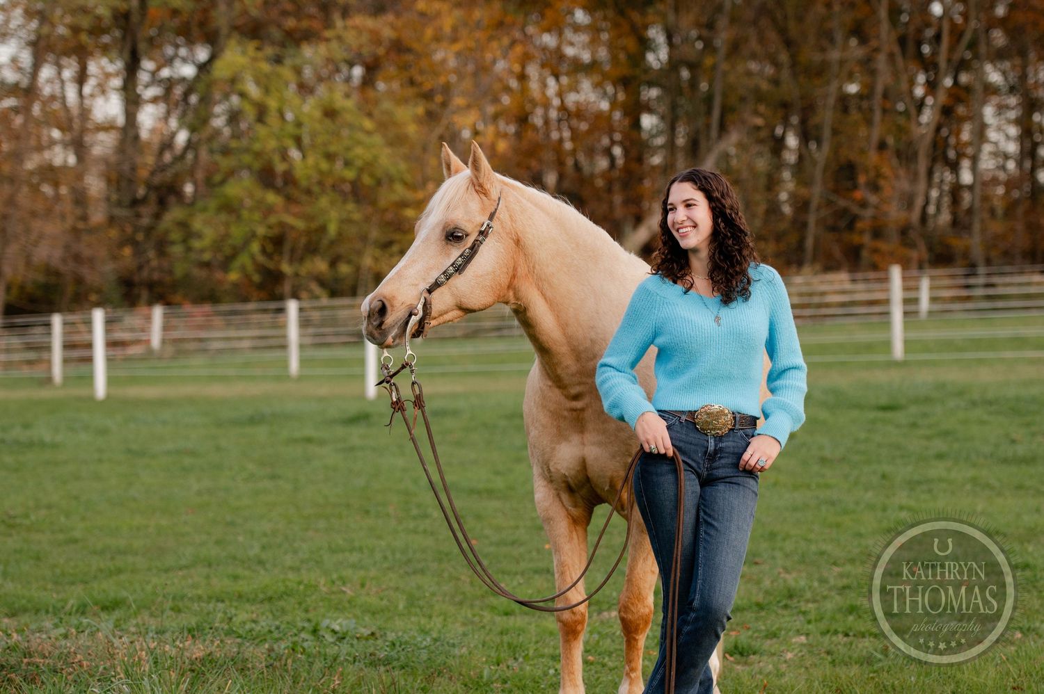 A golden palomino horse stands in a green pasture with white fencing and fall trees in background.
