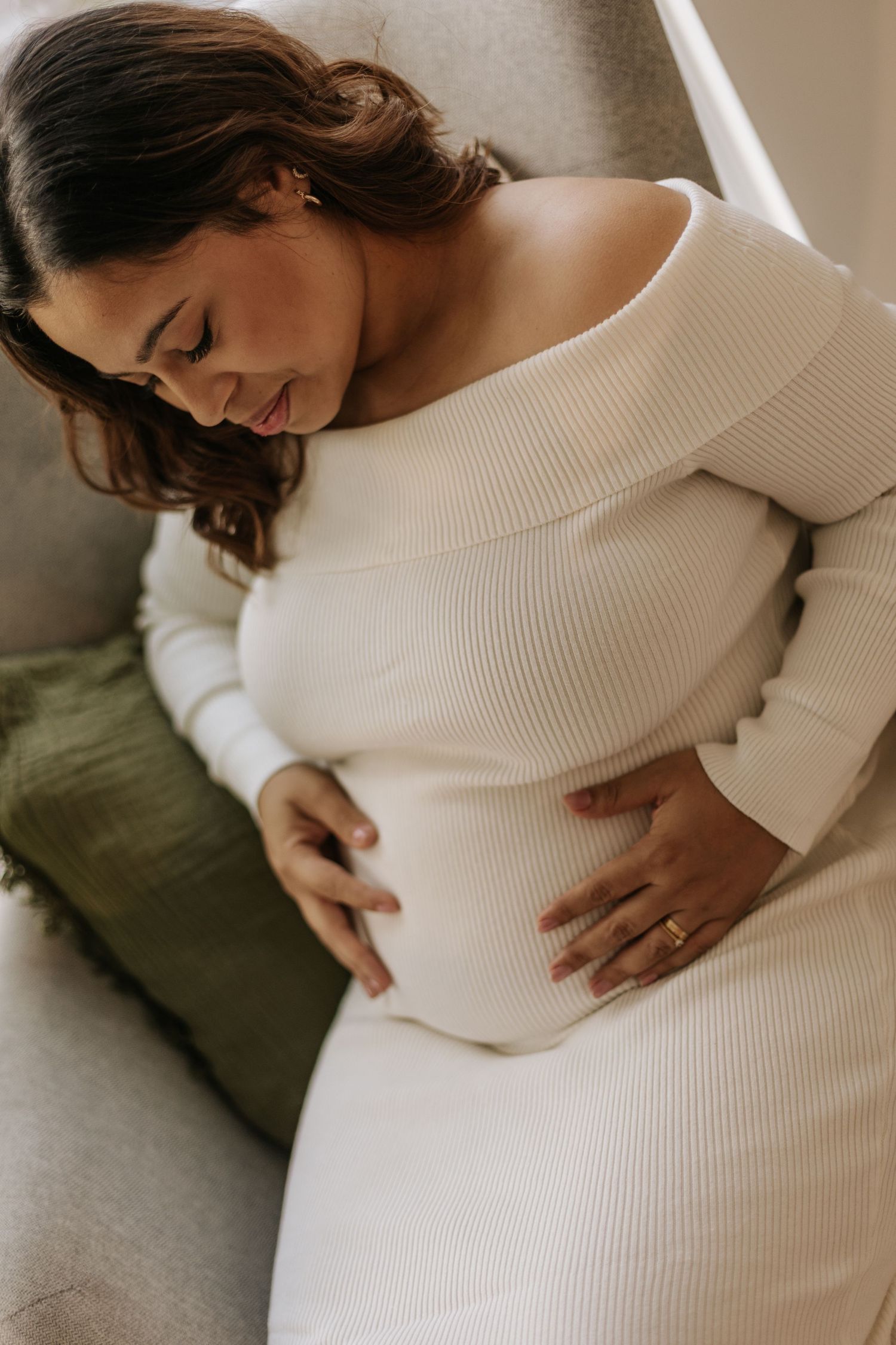 pregnant woman sitting in her armchair, looking down at her bell, wearing a white dress
