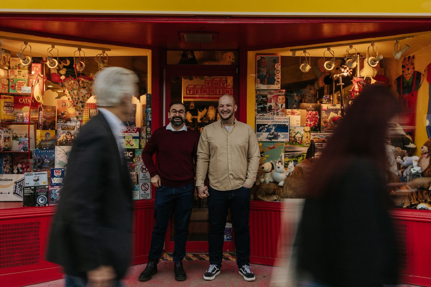 male couple holding hands in front of a toy store while people walk in front of them