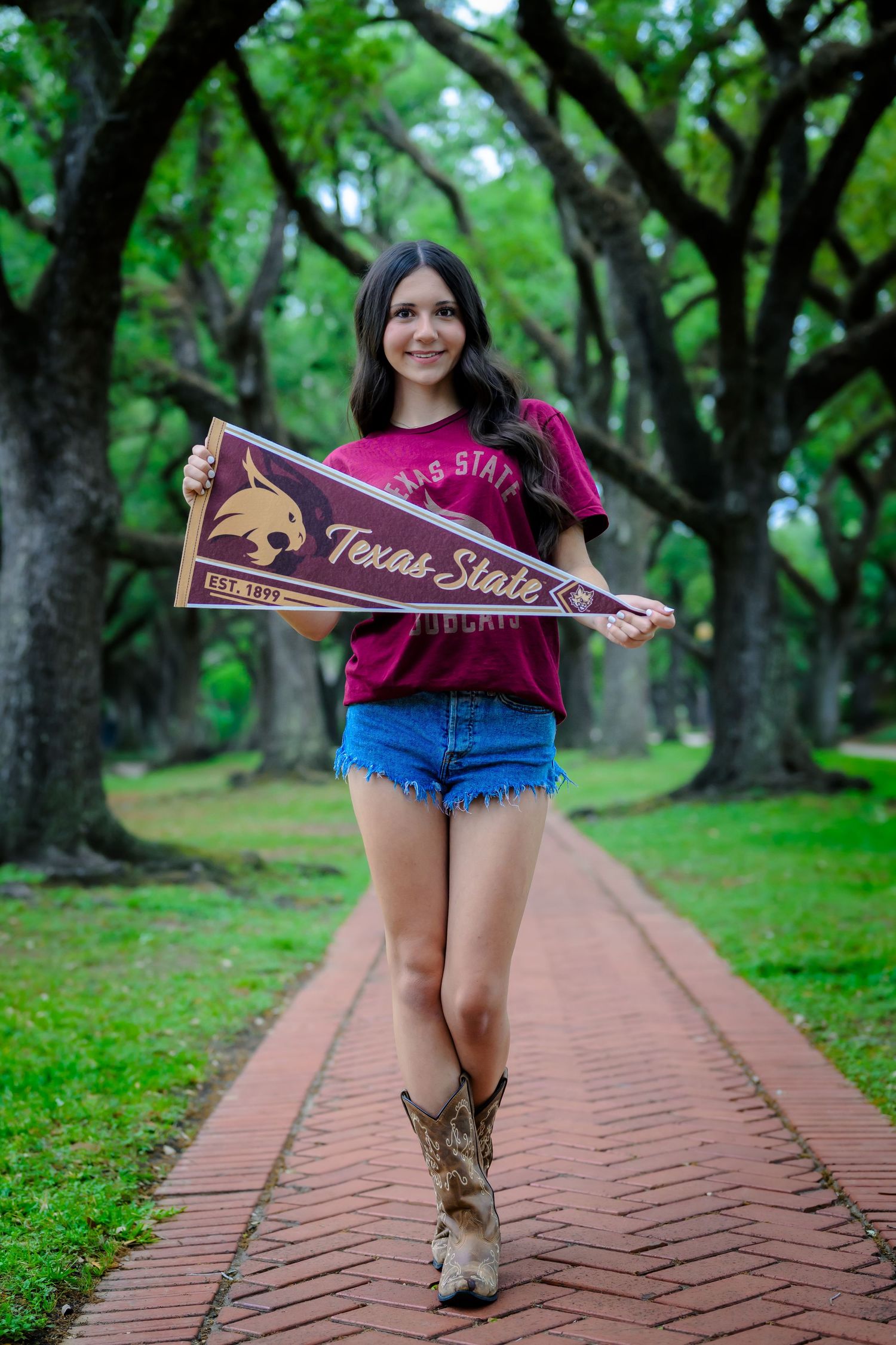 Student in cowboy boots holds Texas State University pennant on brick pathway surrounded by green trees.