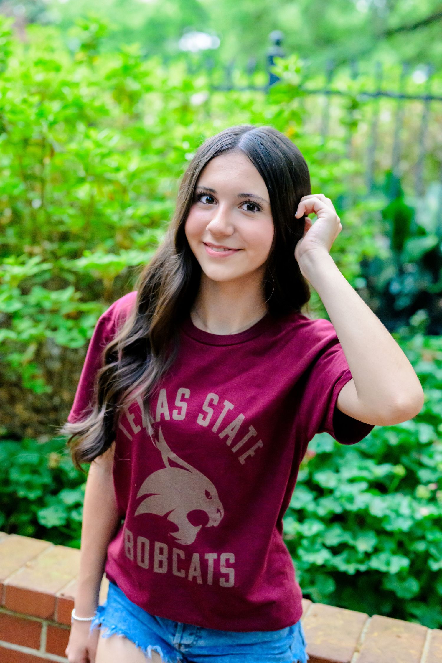 A student wearing a Texas State Bobcats maroon t-shirt poses in front of greenery on a brick wall ledge.