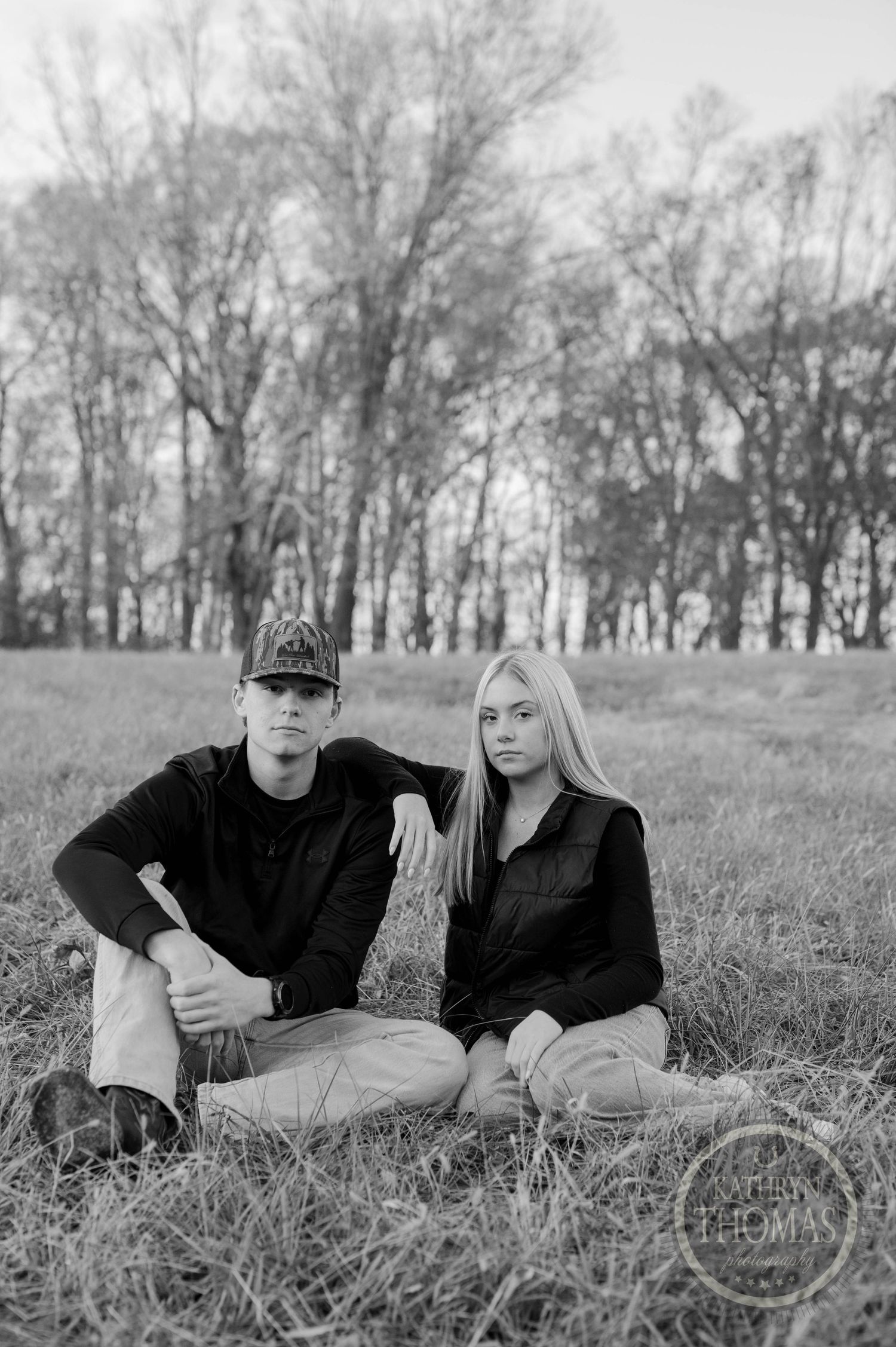 Black and white portrait of siblings sitting in field with bare trees in background.