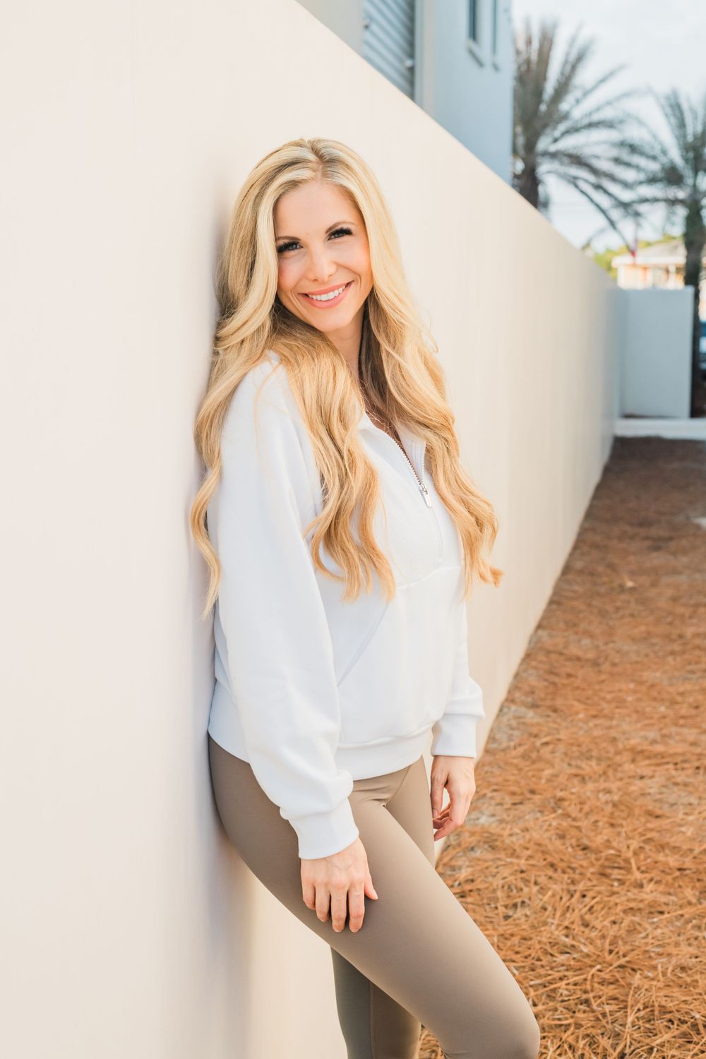 Casual outdoor portrait against white wall featuring flowing blonde hair, white sweater and beige pants.