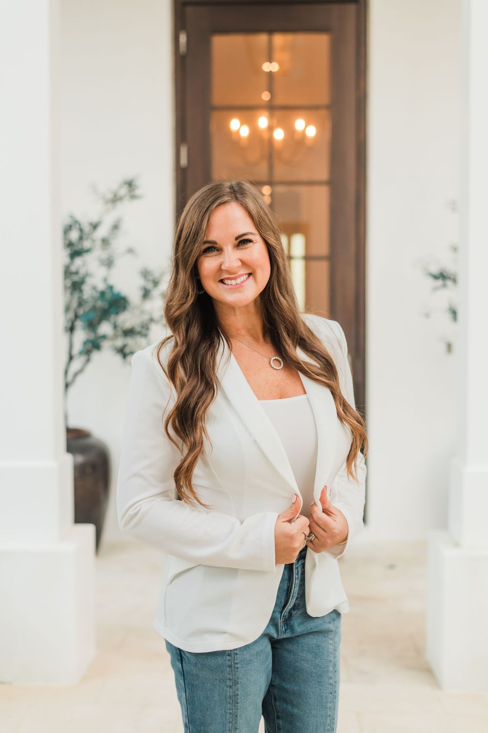 A woman in a white blazer and jeans smiles while standing in front of an elegant entryway with a chandelier.