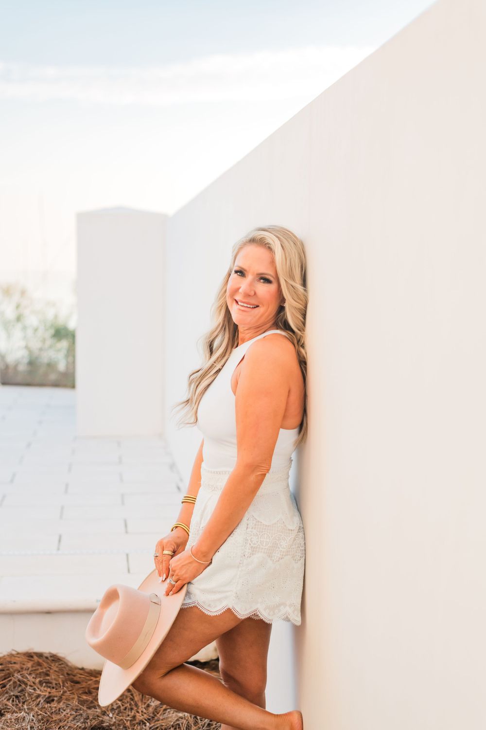A person in an orange cardigan and white dress poses casually against a white wall while holding a tan hat.
