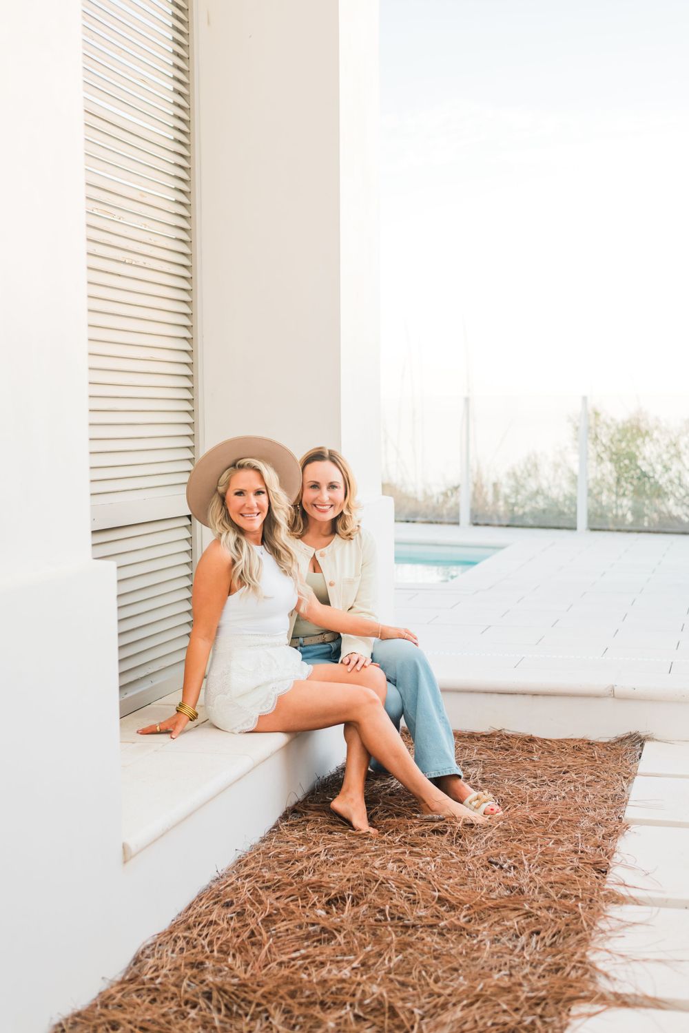 Two women in casual summer outfits sitting on a white ledge by a pool with mulch landscaping in the foreground.
