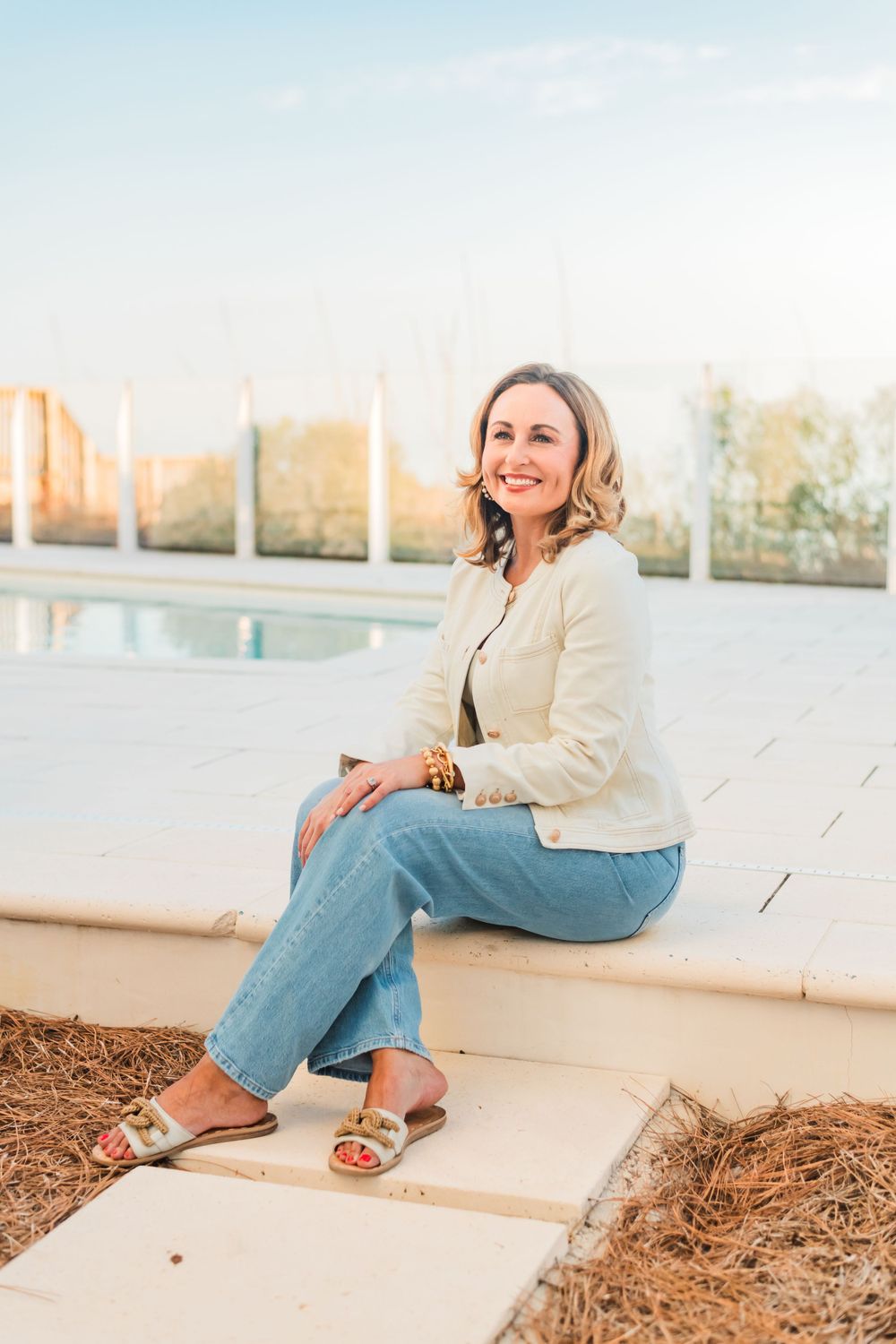 Woman in cream blazer and jeans sits casually outdoors by pool during golden hour sunset.