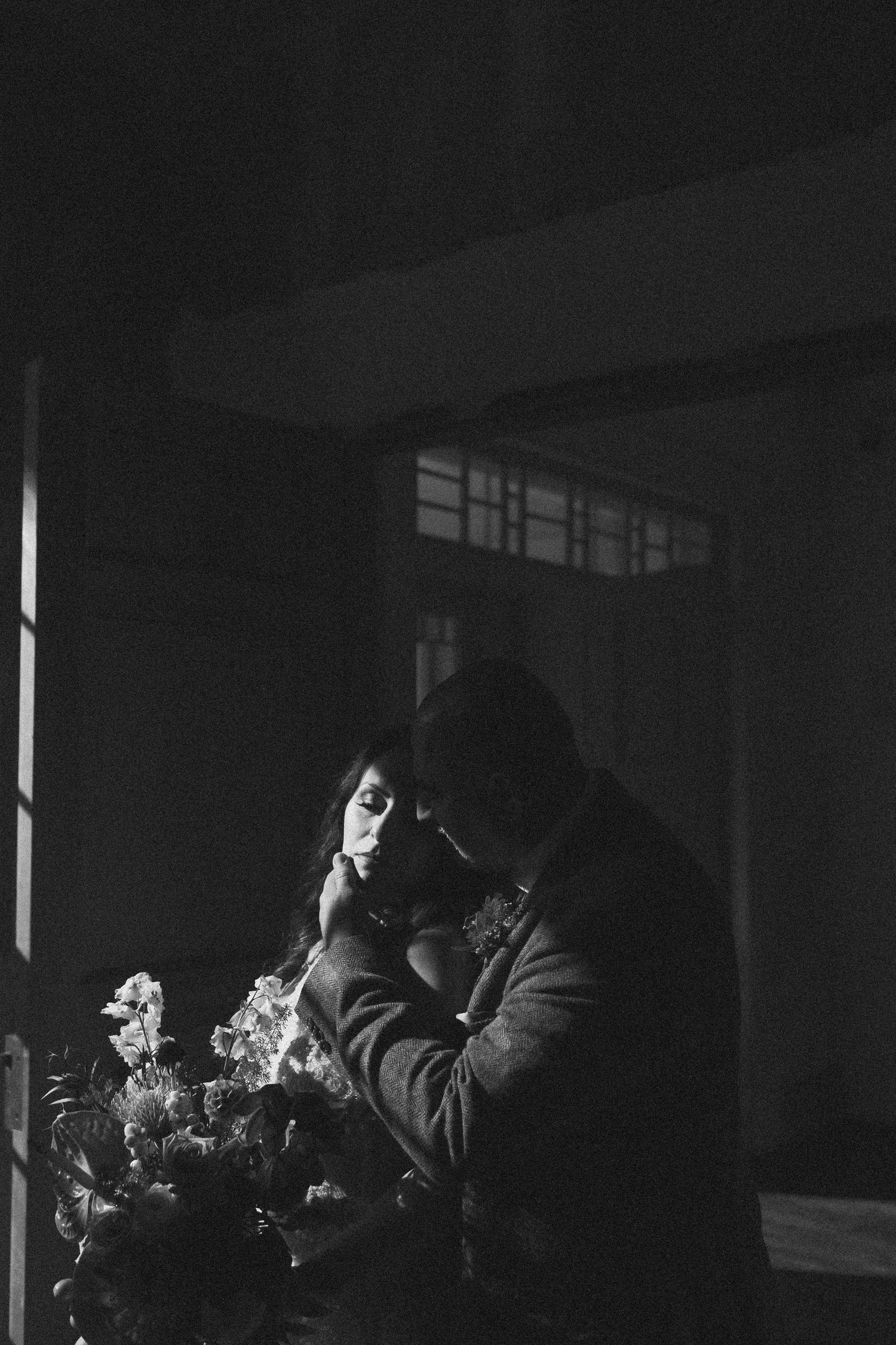 A dark atmospheric noir scene shows a silhouetted figure in a coat standing next to flowers by a window.