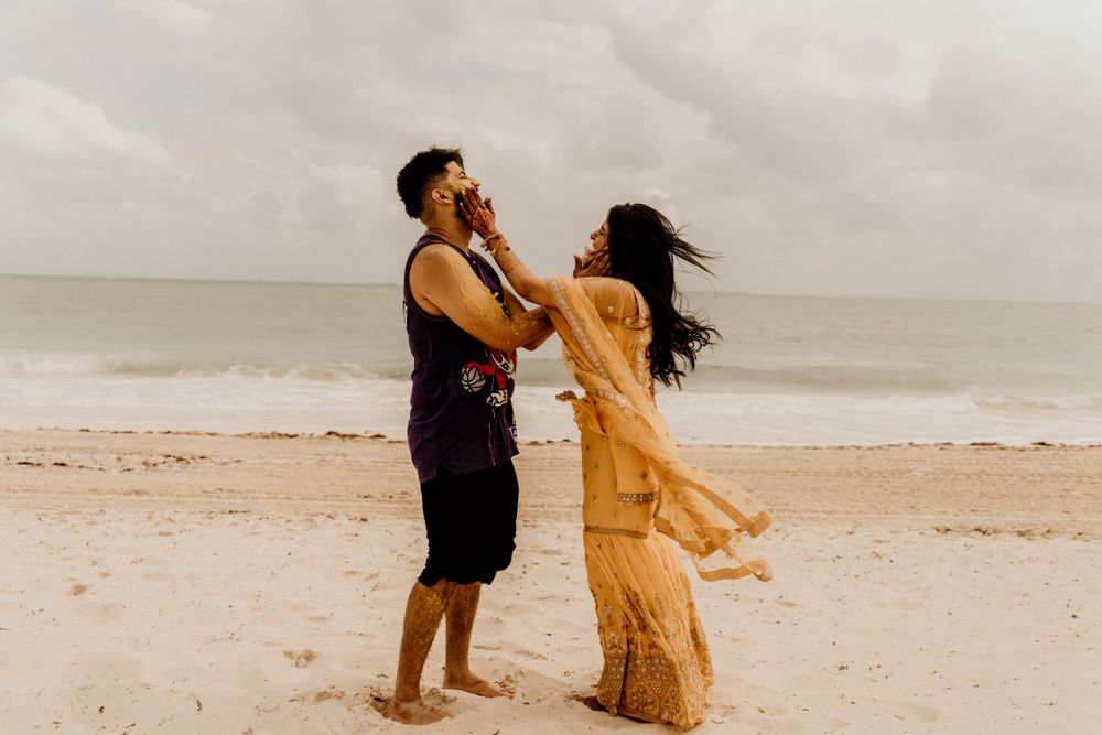 A couple dances playfully together on a cloudy beach, her dress flowing in the wind as they embrace.