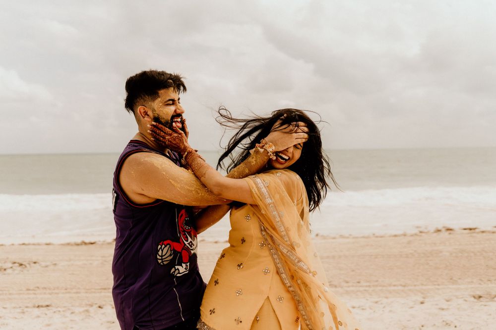 A couple dances together on the beach, spinning playfully with wind-swept hair against an overcast sky.
