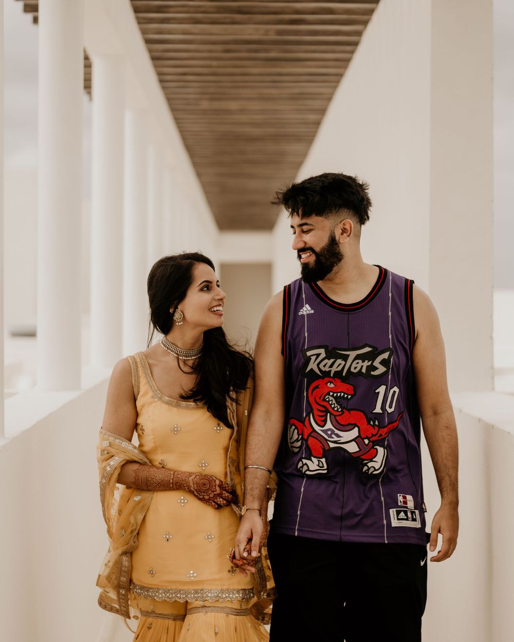 A couple in traditional Indian dress and basketball jersey holding hands in a modern hallway with wooden beams.