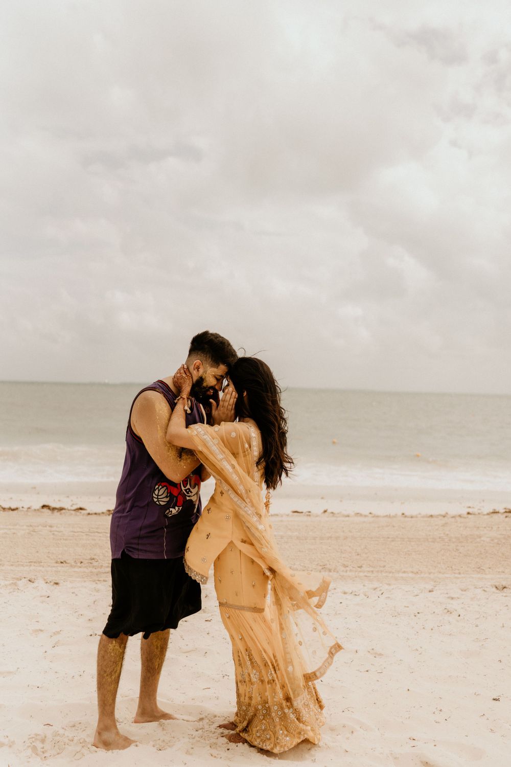 A romantic couple embraces on a sandy beach with cloudy skies and ocean waves in the background.
