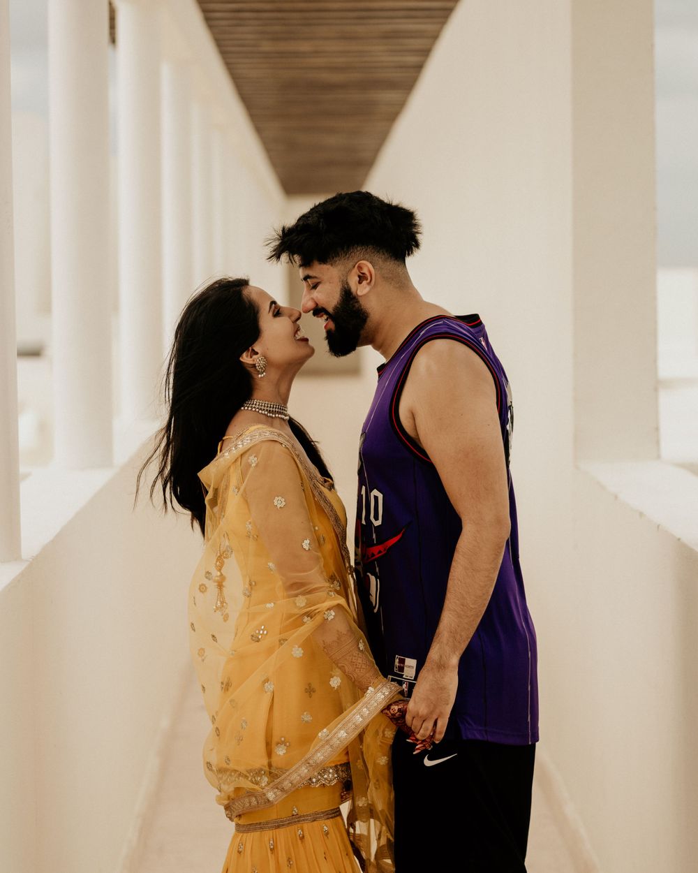 A couple shares an intimate moment in a long hallway, with her wearing a yellow traditional dress and him in a purple tank top.