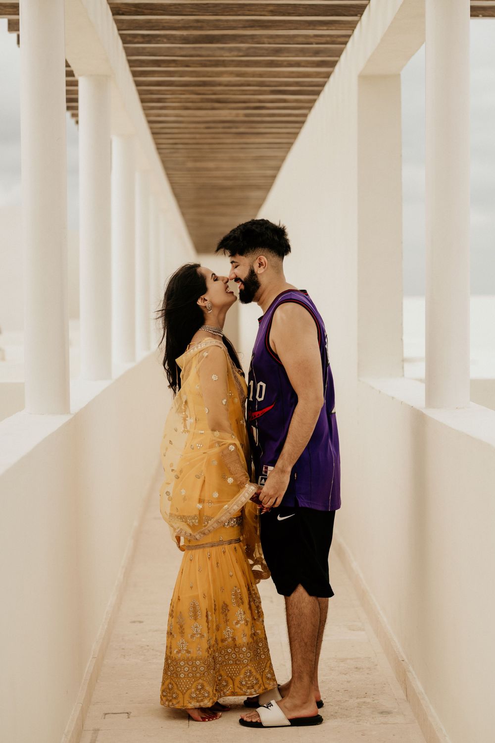 Couple sharing an intimate moment in a white pillared hallway, with her in yellow traditional dress and him in purple jersey.