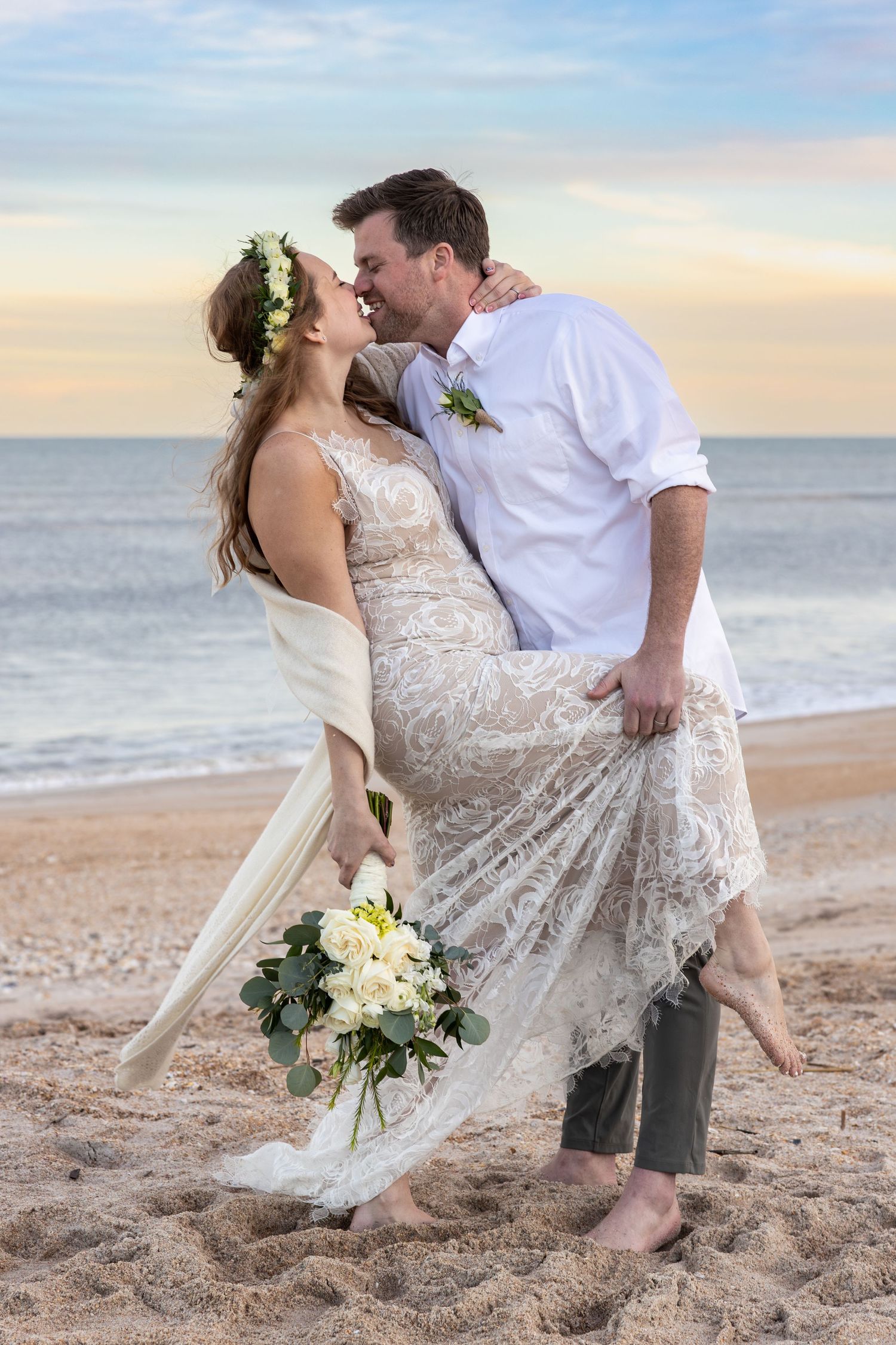Romantic beach wedding portrait with flowing lace dress against sunset sky.