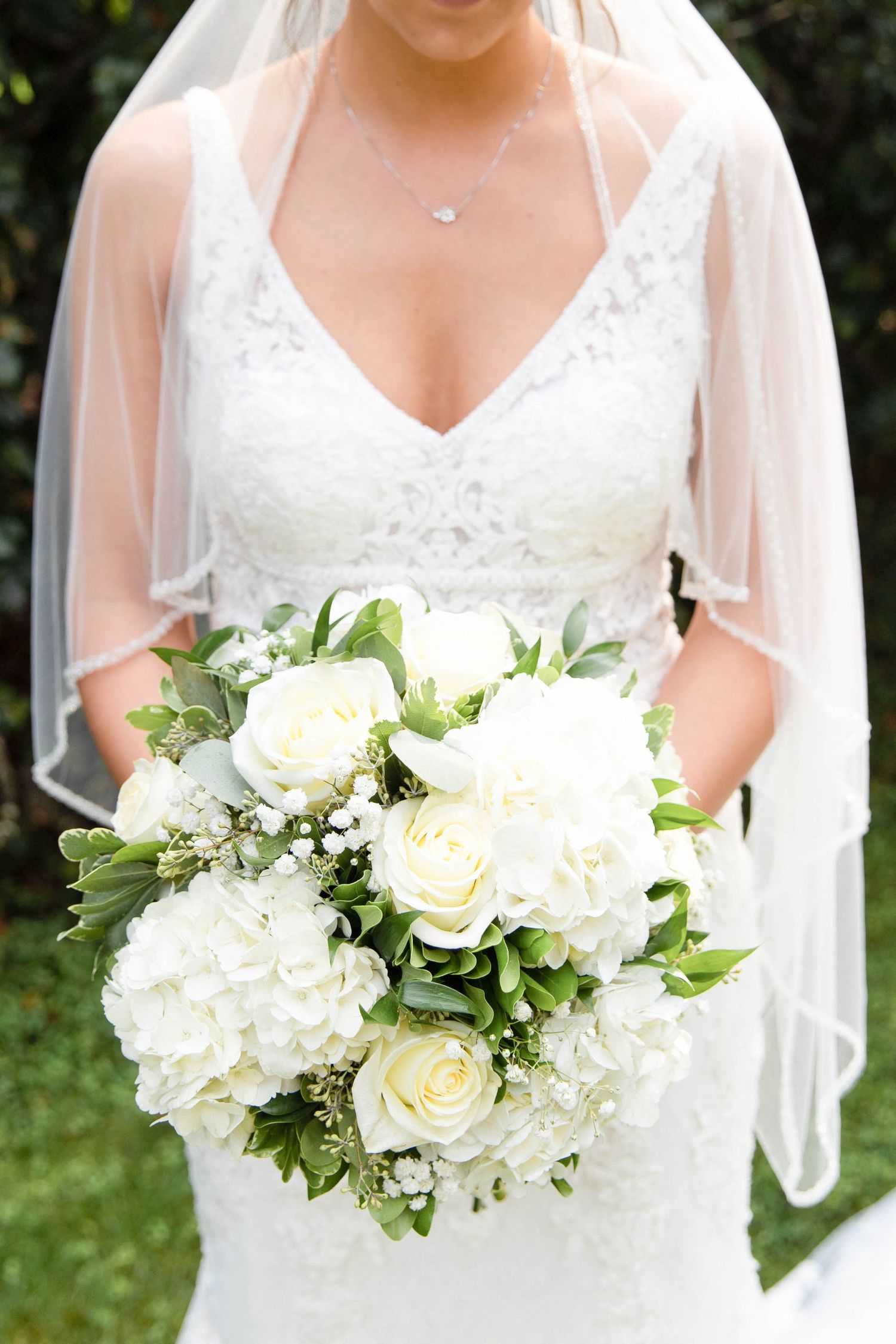 Beautiful classic white bridal bouquet with roses, hydrangeas and greenery held against a wedding dress with veil.