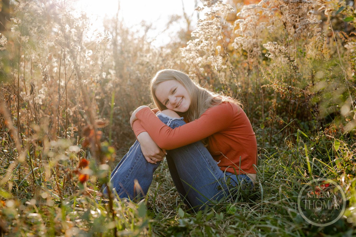 Candid outdoor photo of someone in orange sweater hugging their knees among fall foliage.