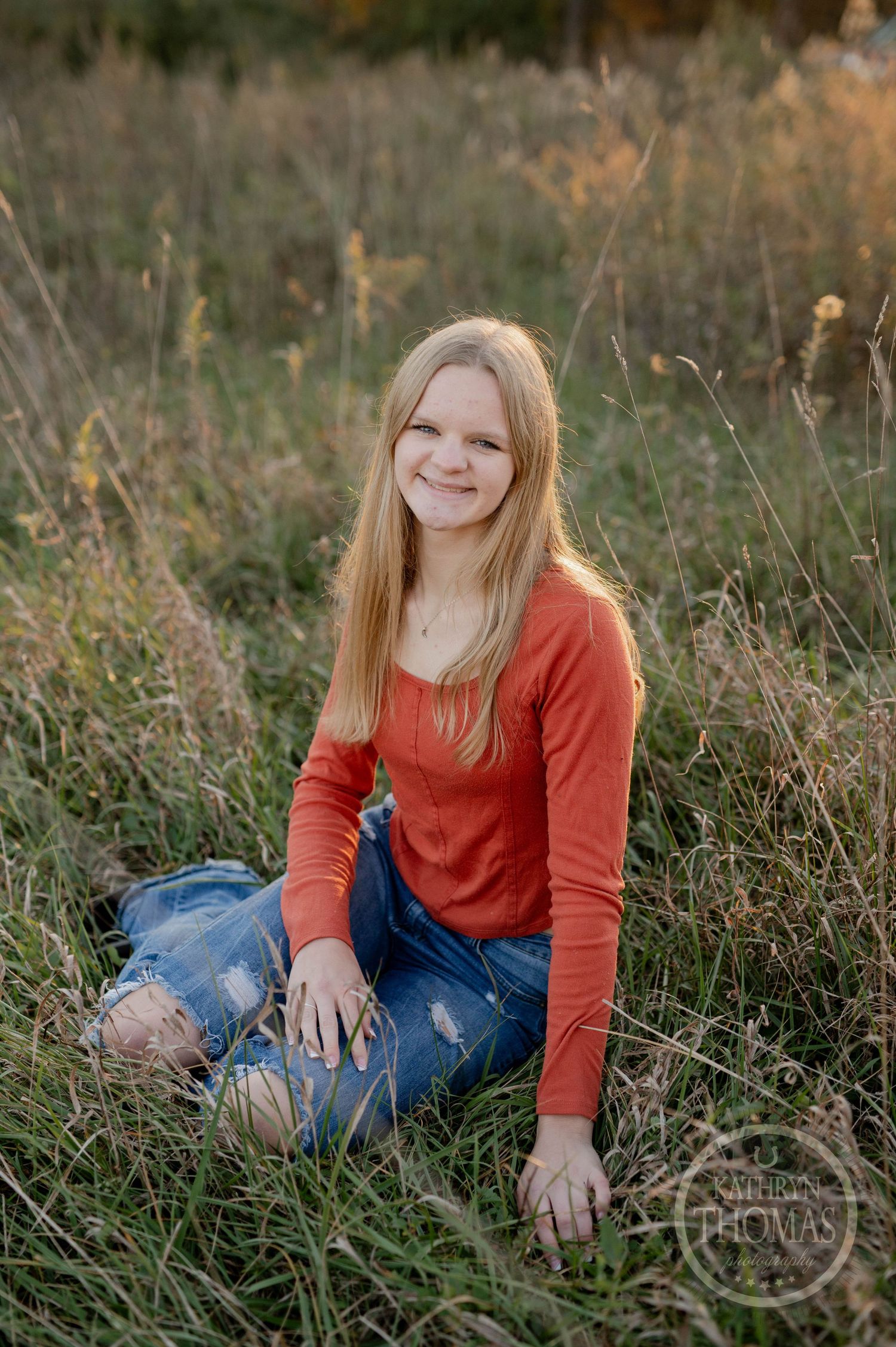 Person in orange top sits cross-legged in autumn field with wild grass and flowers.