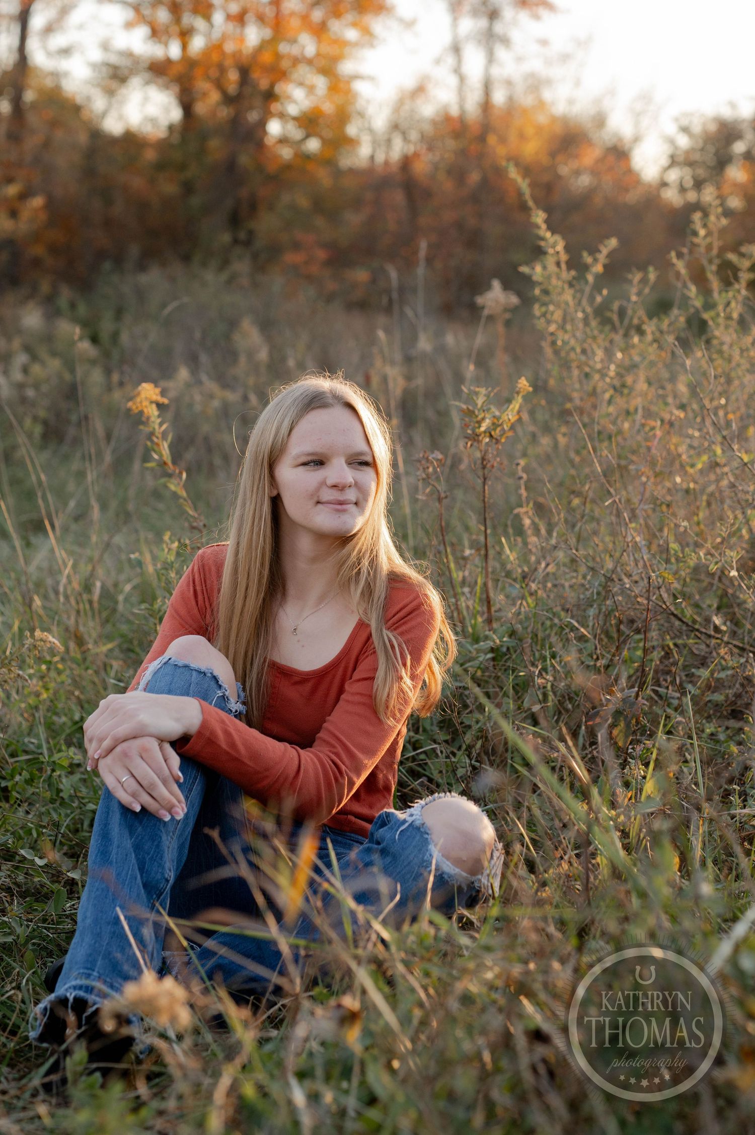A person in rust-colored sweater relaxes in tall grass during golden hour with fall foliage in background.