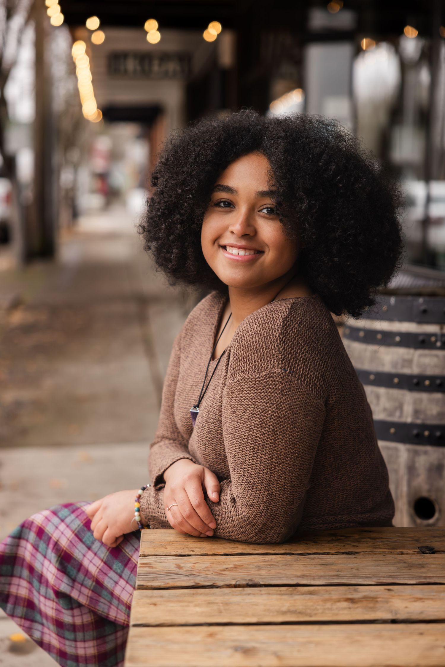 A smiling individual with natural curly hair wearing a brown sweater sits at a wooden table outdoors.