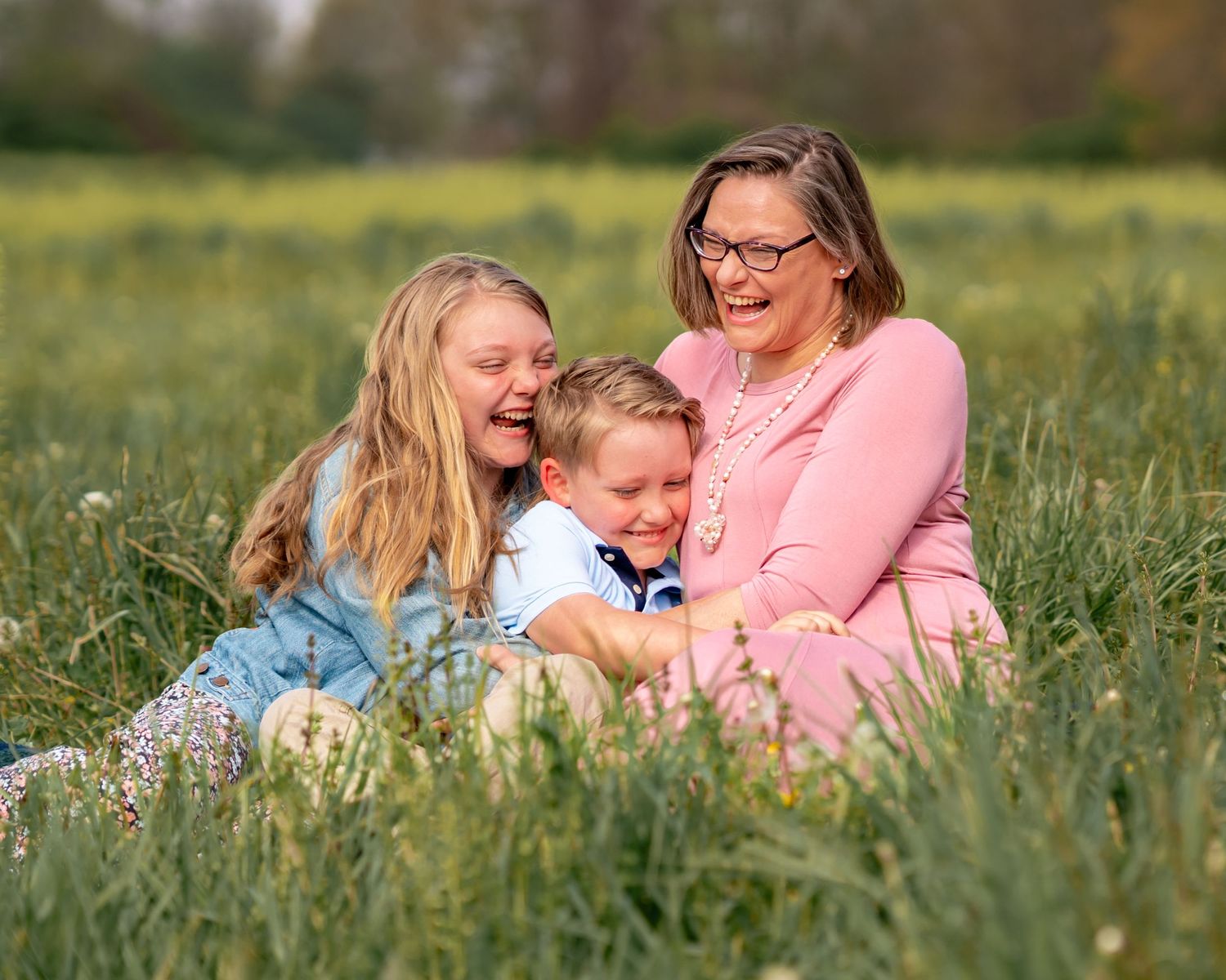 Three people sharing joyful laughter while sitting together in a sunny spring meadow with tall green grass and flowers.