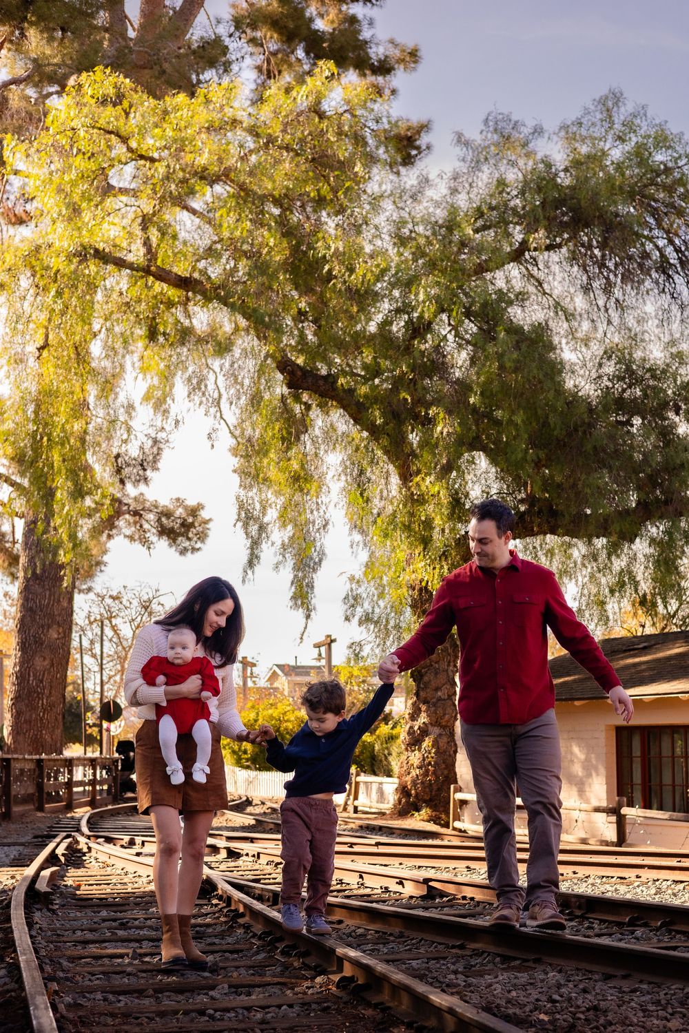 Family walking along train tracks at Old Poway Park, mom holding baby girl, both parents holding little boy’s hands during holiday session.