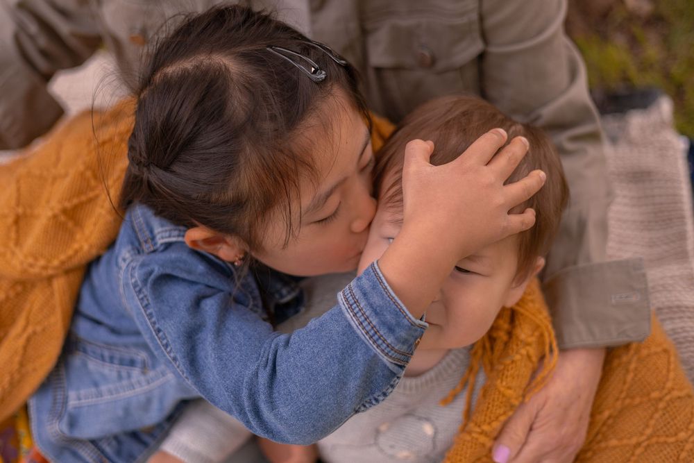 Little girl kissing baby cousin while gently holding his head still — candid, nostalgic family moment at Old Poway Park in Poway, CA.