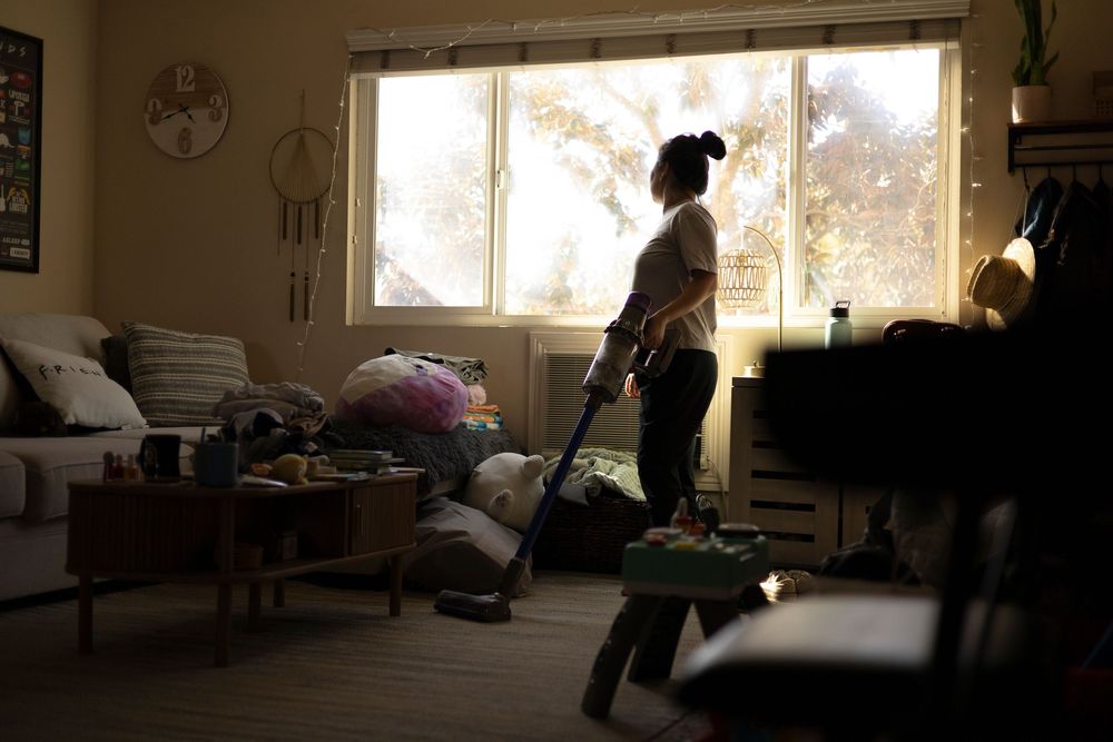 Mom holding a vacuum, looking out a bright window with the rest of the frame dark and moody — nostalgic, introspective moment.