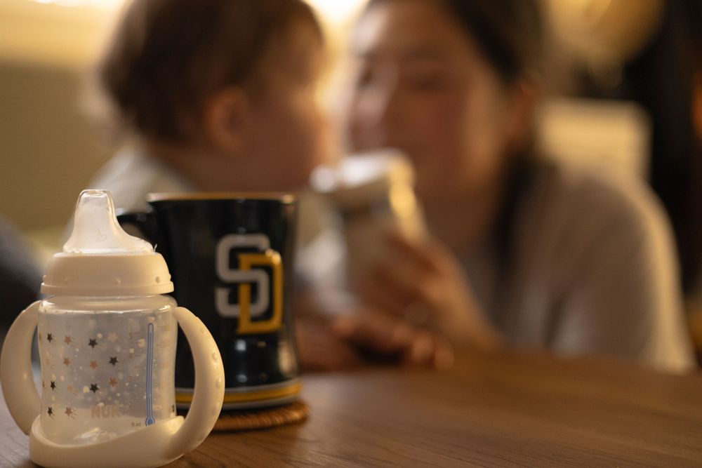 Baby bottle and coffee mug on the coffee table and mom giving water cup to baby boy in a softly blurred background in Rancho Penasquitos.