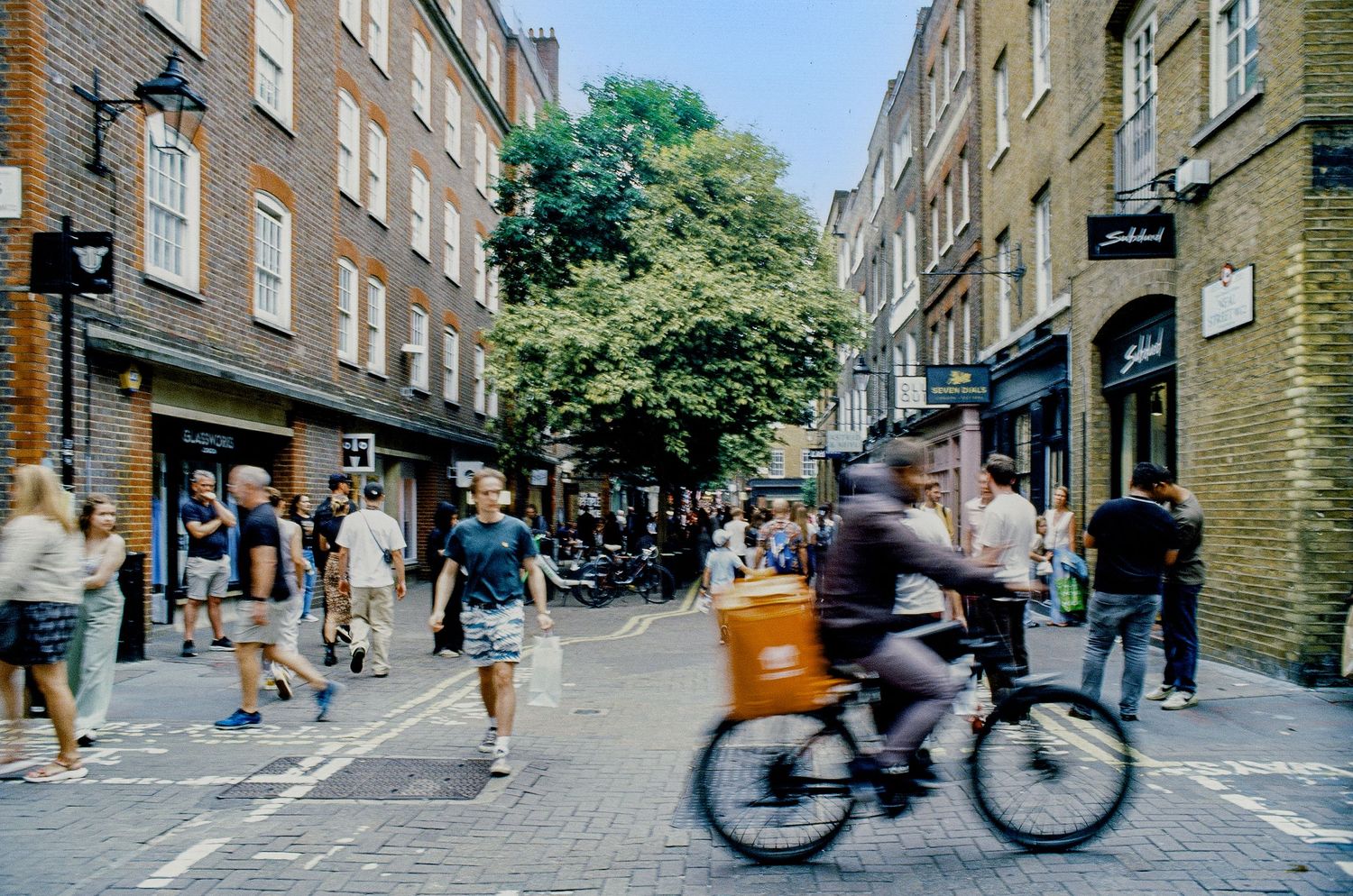 Cyclist and pedestrians navigate a busy cobblestone street lined with shops and historic brick buildings in London.