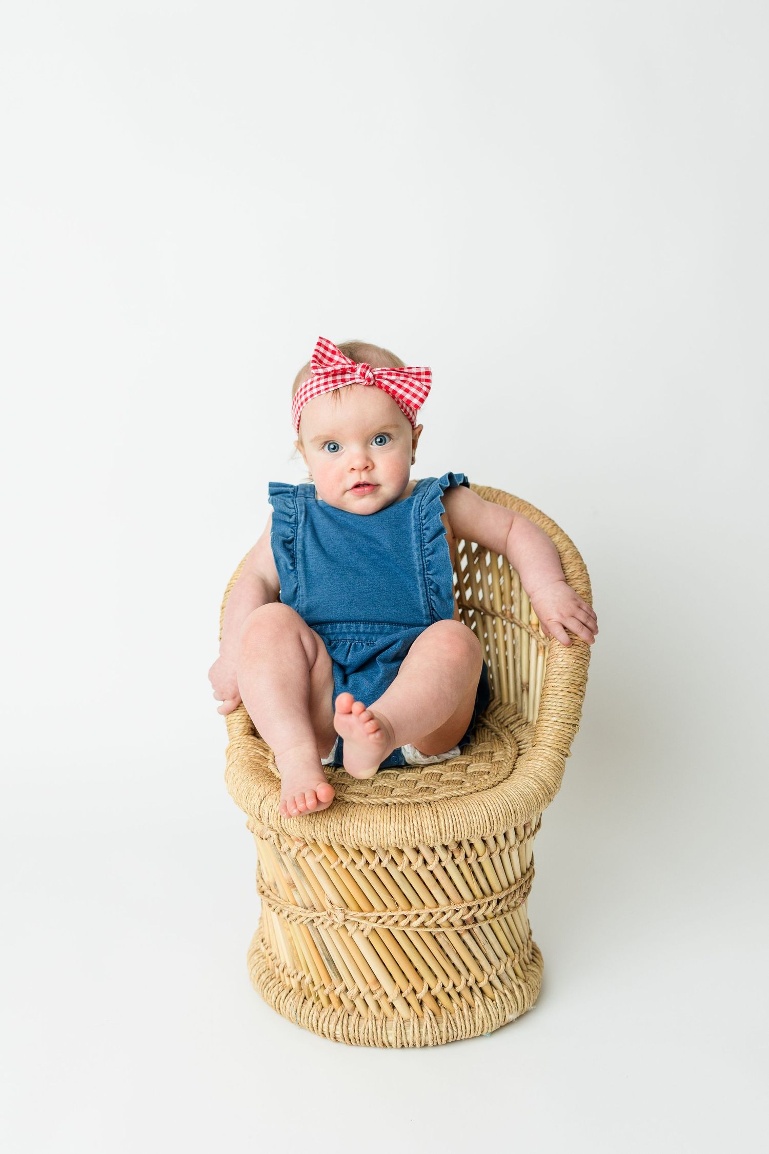 Small baby wearing blue denim romper and red bow sits in woven rattan peacock chair against white background.