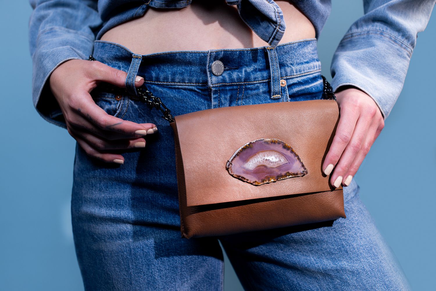 A brown leather purse with a metallic lip design sits against denim jeans on a blue background.