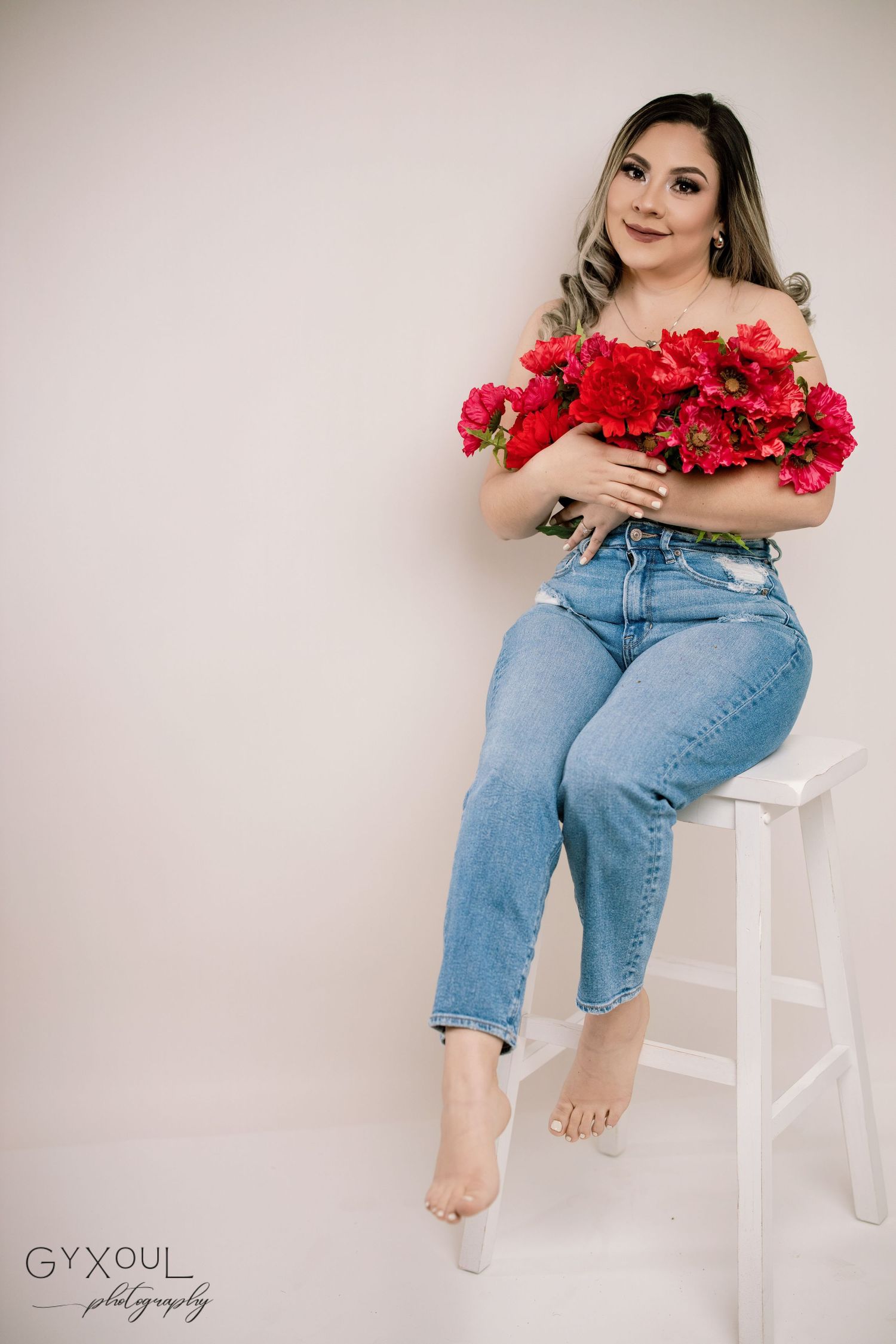 A casual portrait with red flowers and light blue jeans on a white stool against a neutral background.
