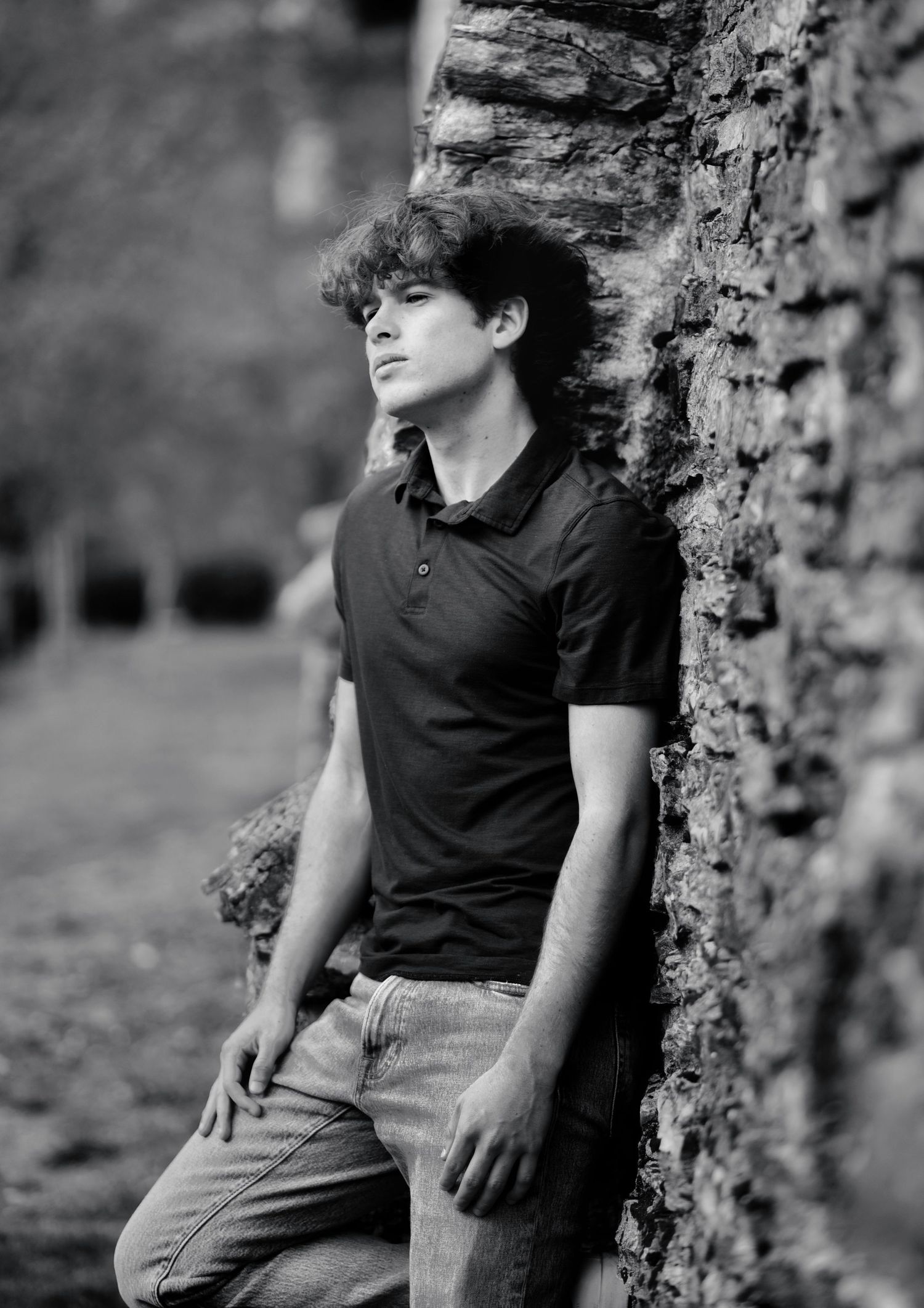 A black and white artistic portrait of a young person in a polo shirt leaning against a stone wall outdoors.