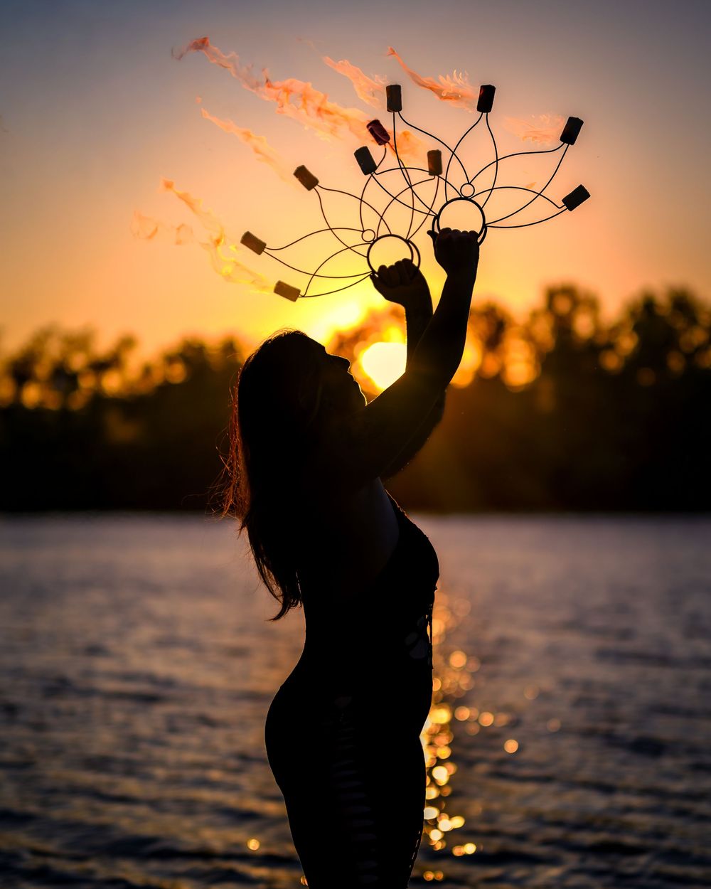 Silhouette of a woman holding a fire fan prop against a golden sunset over a lake.