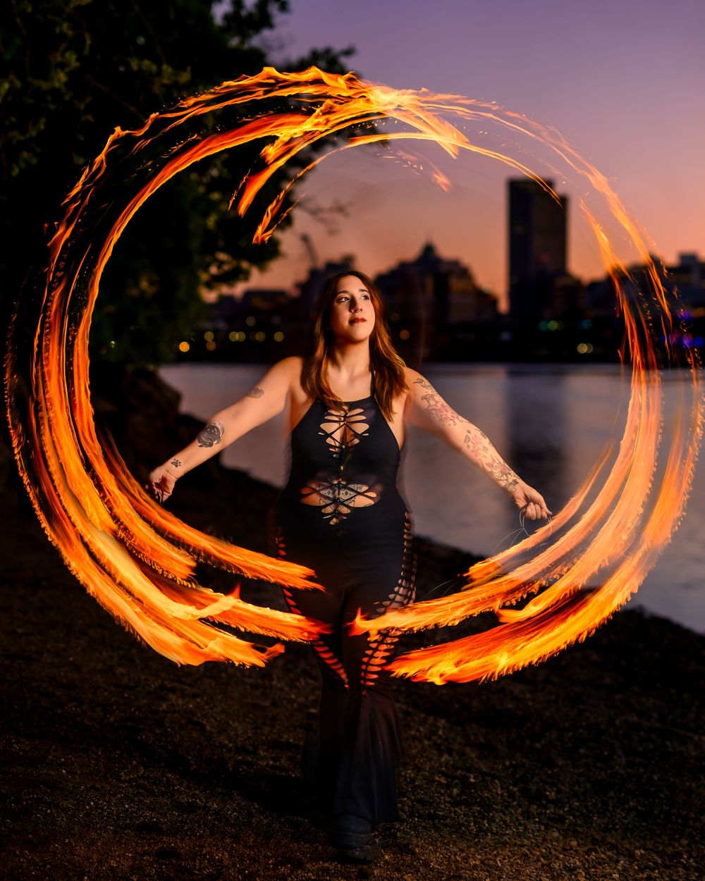 Woman performing fire dancing at dusk, creating a glowing circular trail of flames near a city waterfront.