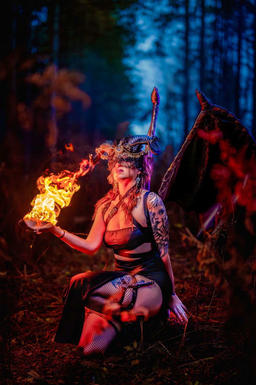 A fire performer in gothic attire kneels in a misty forest setting with dramatic blue lighting and flames.