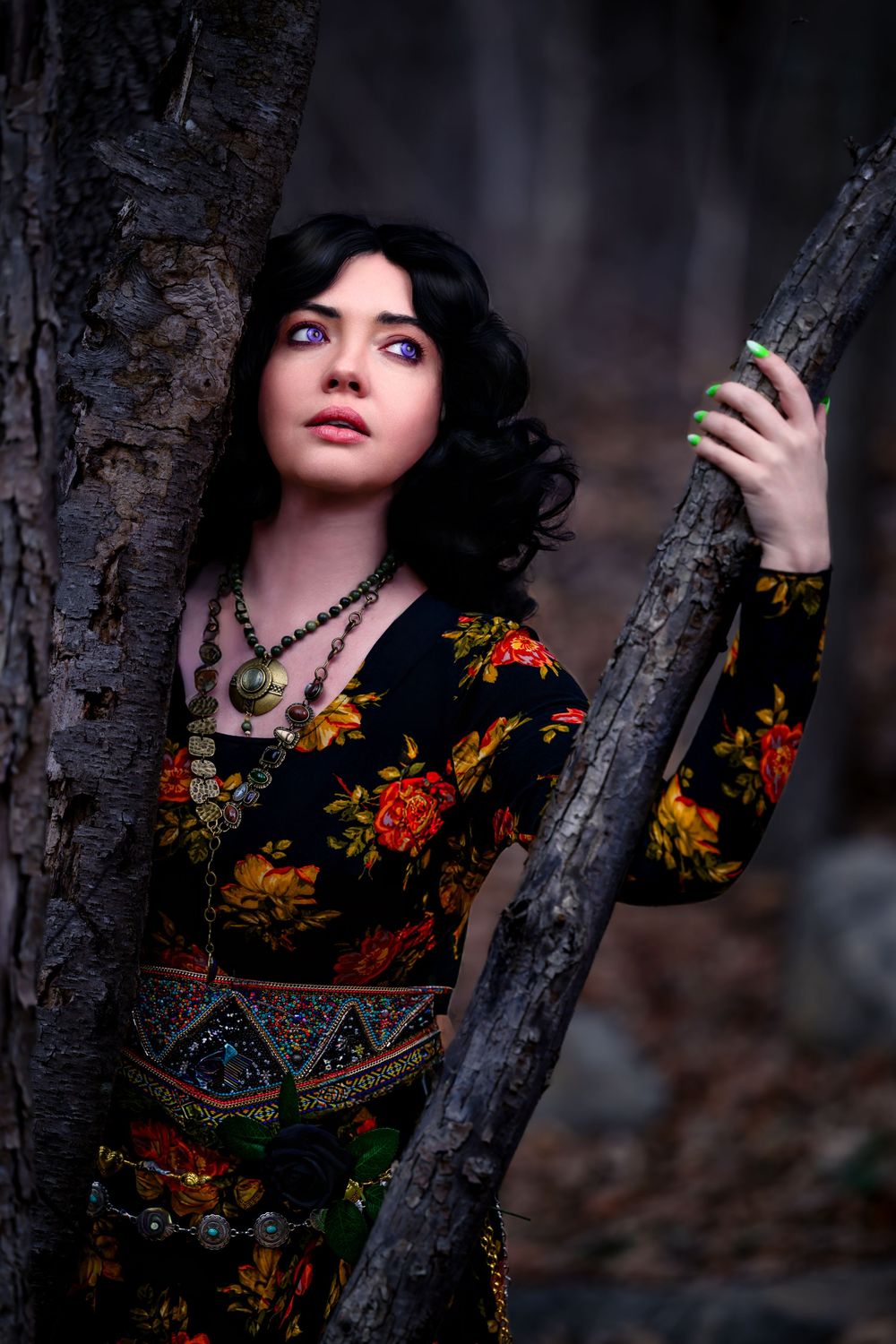 Young woman with dark hair and blue eyes in floral dress leaning against trees in a dark forest setting.