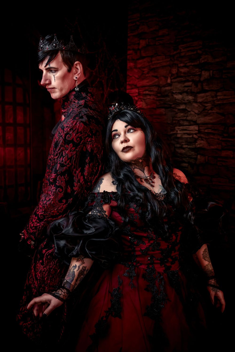 Gothic couple in red and black Victorian attire with crowns pose dramatically against a dark stone wall.