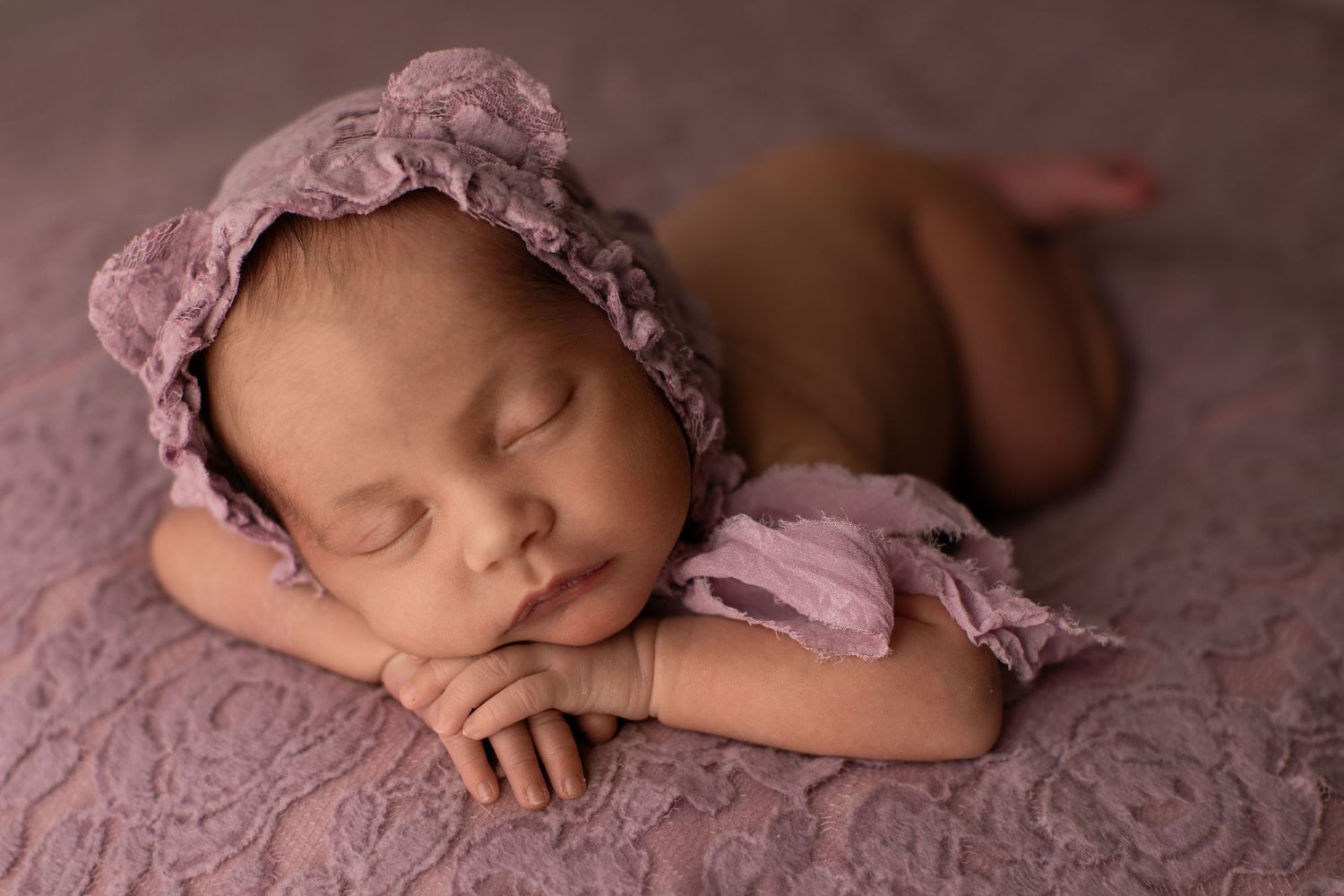 Peaceful sleeping newborn wearing pink lace bonnet rests on dusty rose textured fabric during professional photo session.