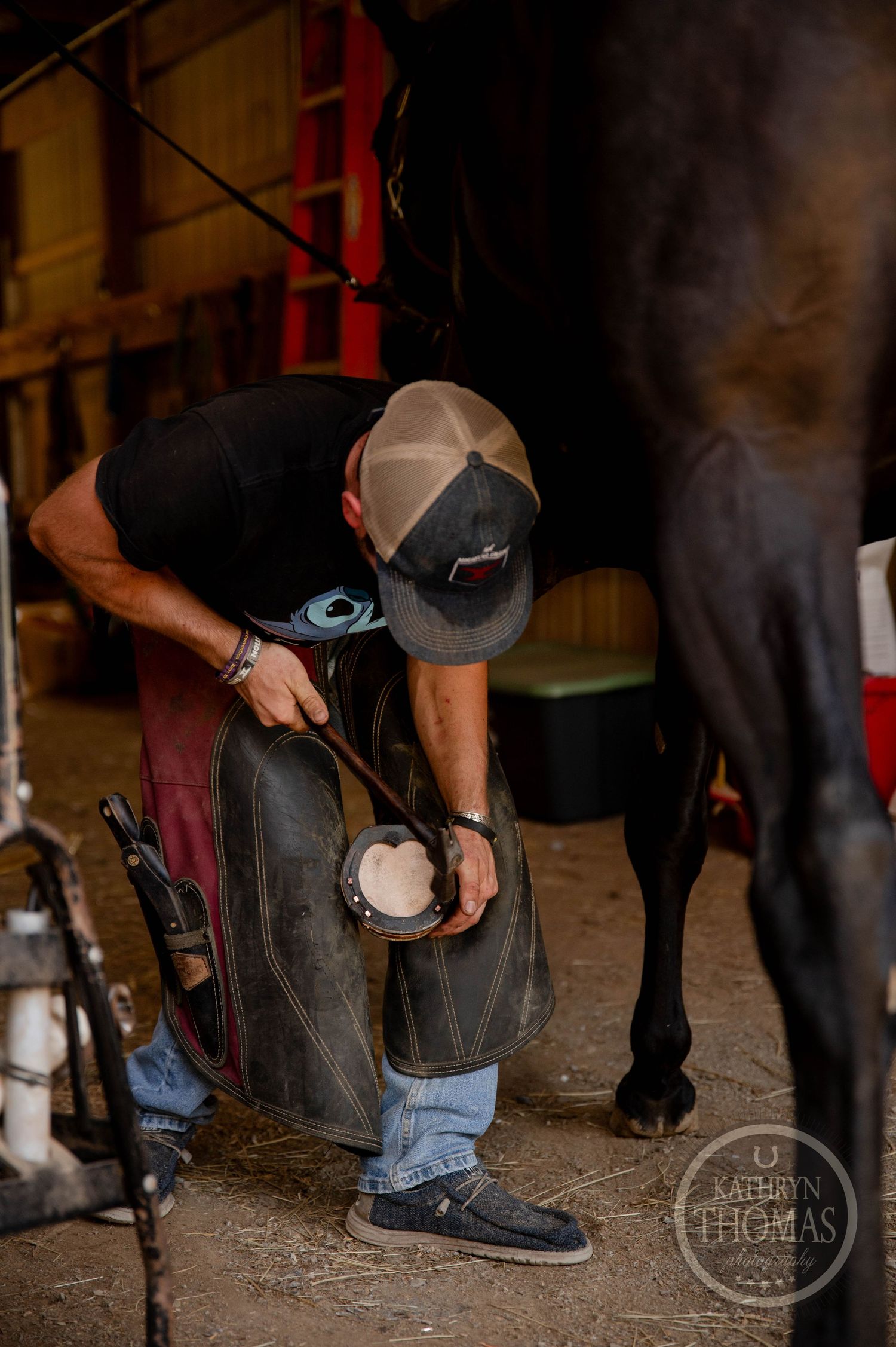 A farrier works on trimming a horse's hoof in a dimly lit stable.