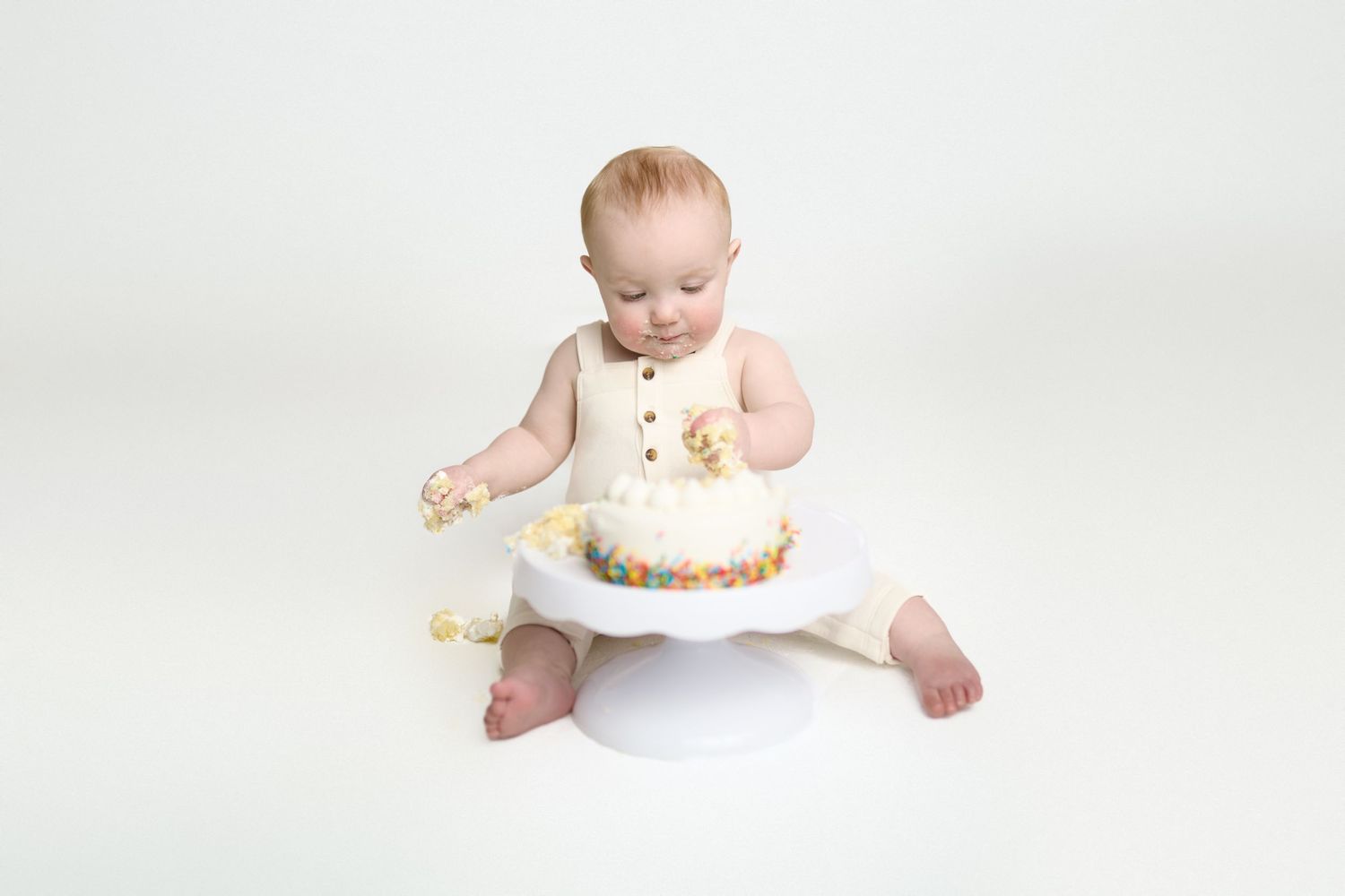 Baby in white outfit sits on floor with birthday cake and frosting on hands against light background.