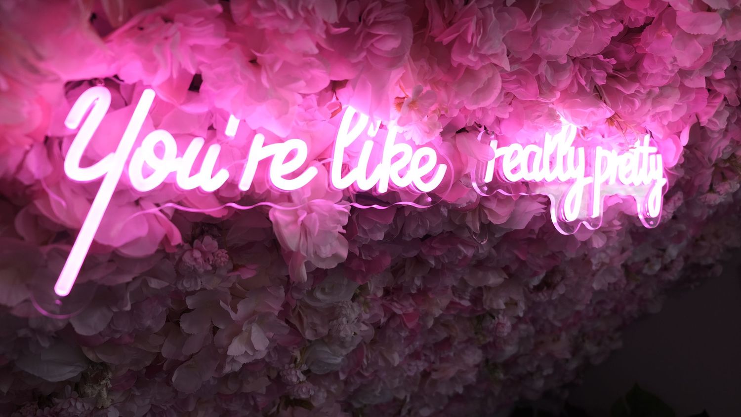 Pink neon sign reading 'You're like, really pretty' mounted on a wall covered with pink floral decorations.