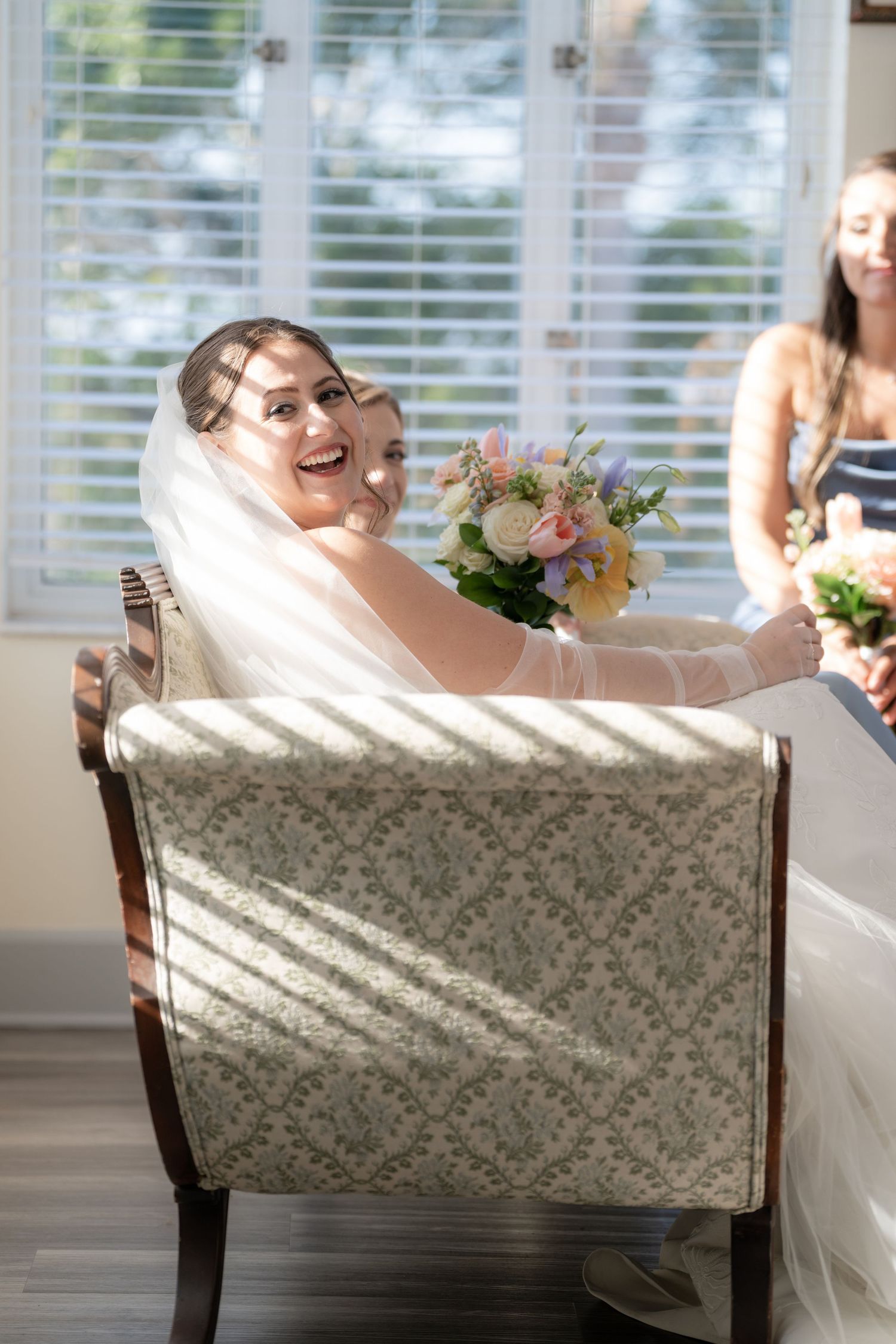 A bride in a white dress laughs joyfully while seated in a patterned armchair near sunlit windows.