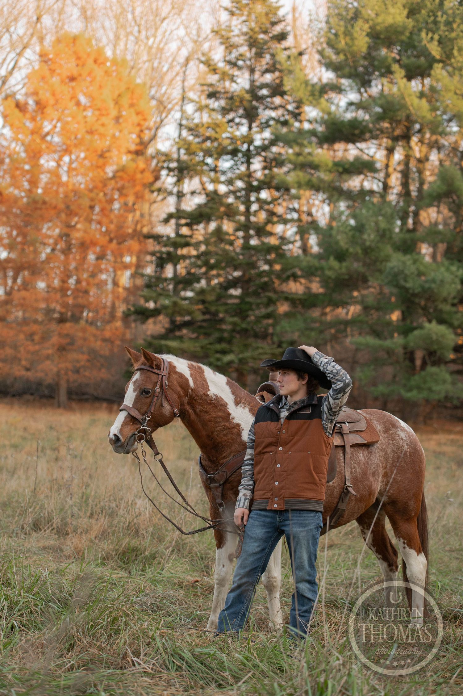 A paint horse with western saddle stands beside a rider in cold weather gear against autumn foliage.