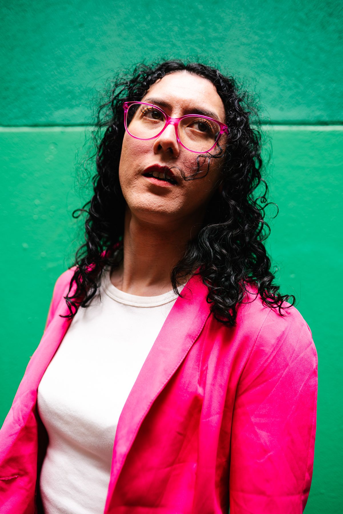 Portrait against green wall featuring pink blazer and glasses with curly hair for an individual branding photoshoot for a Melbourne comedian