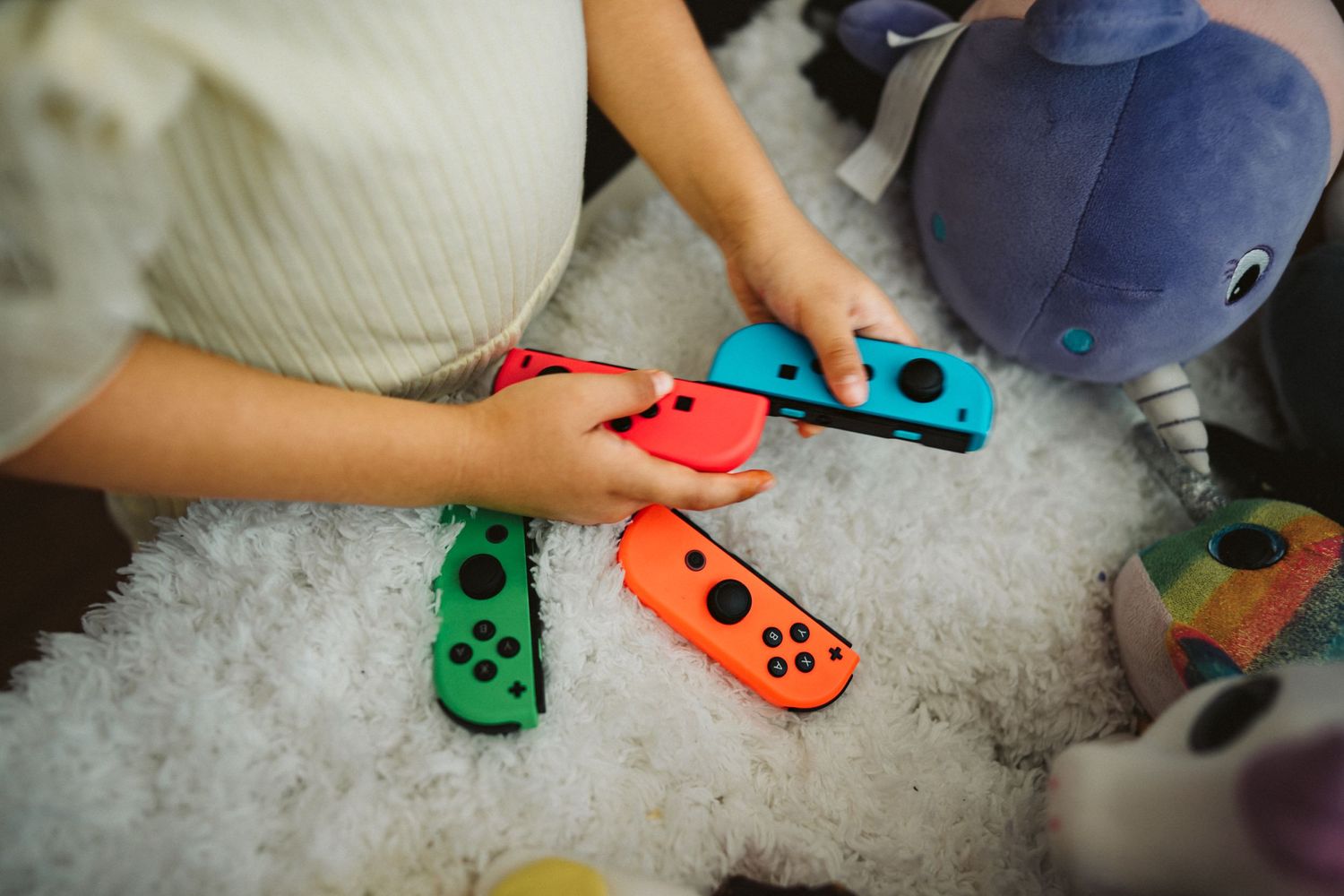 Nintendo Switch Joy-Con controllers in bright colors being held over a white fluffy blanket or carpet.